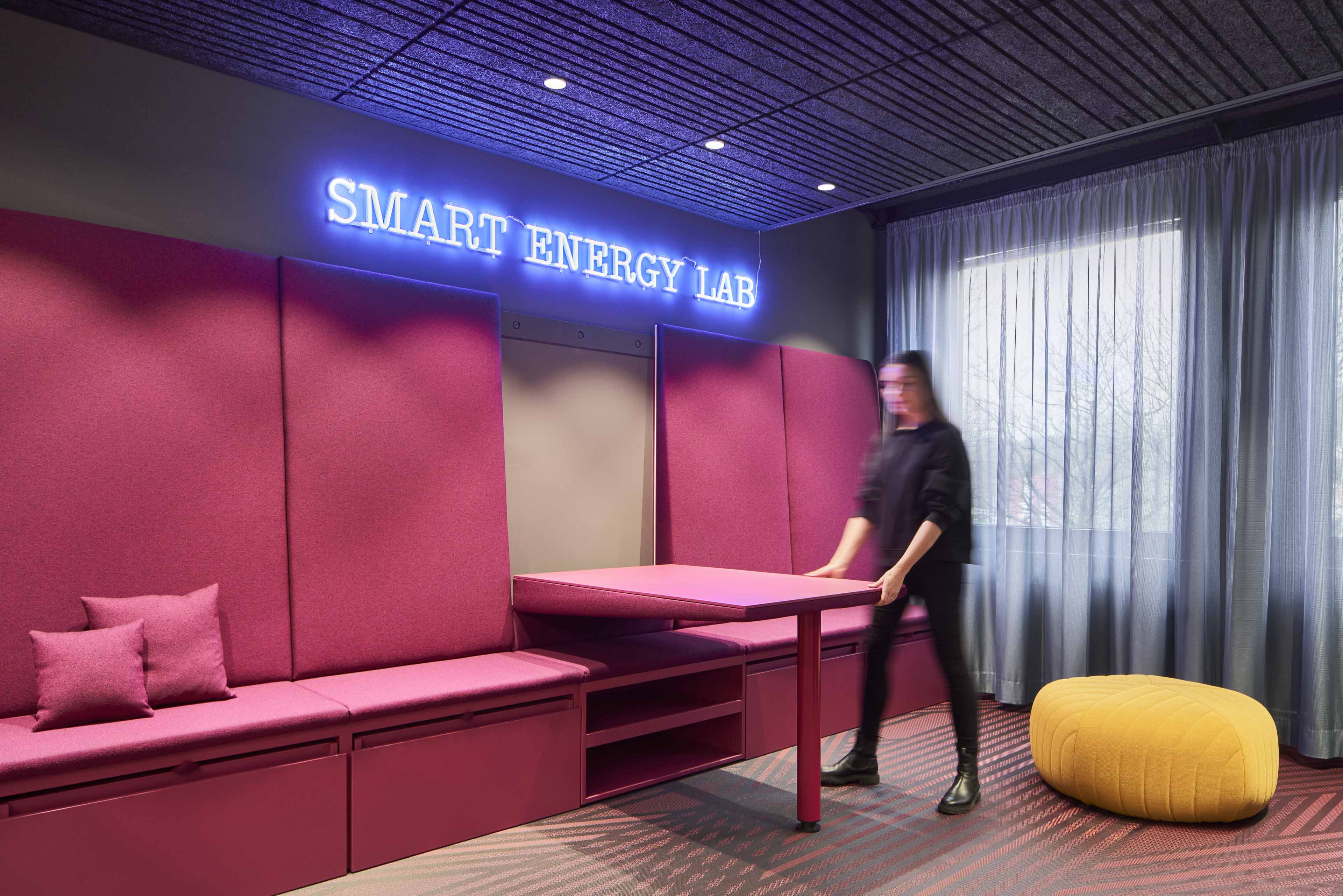 Ein moderner Raum mit einer pinkfarbenen Sitzbank, einem ausklappbaren Tisch, einem gelben Sitzpouf und einem Leuchtschriftzug 'SMART ENERGY LAB' an der Wand.