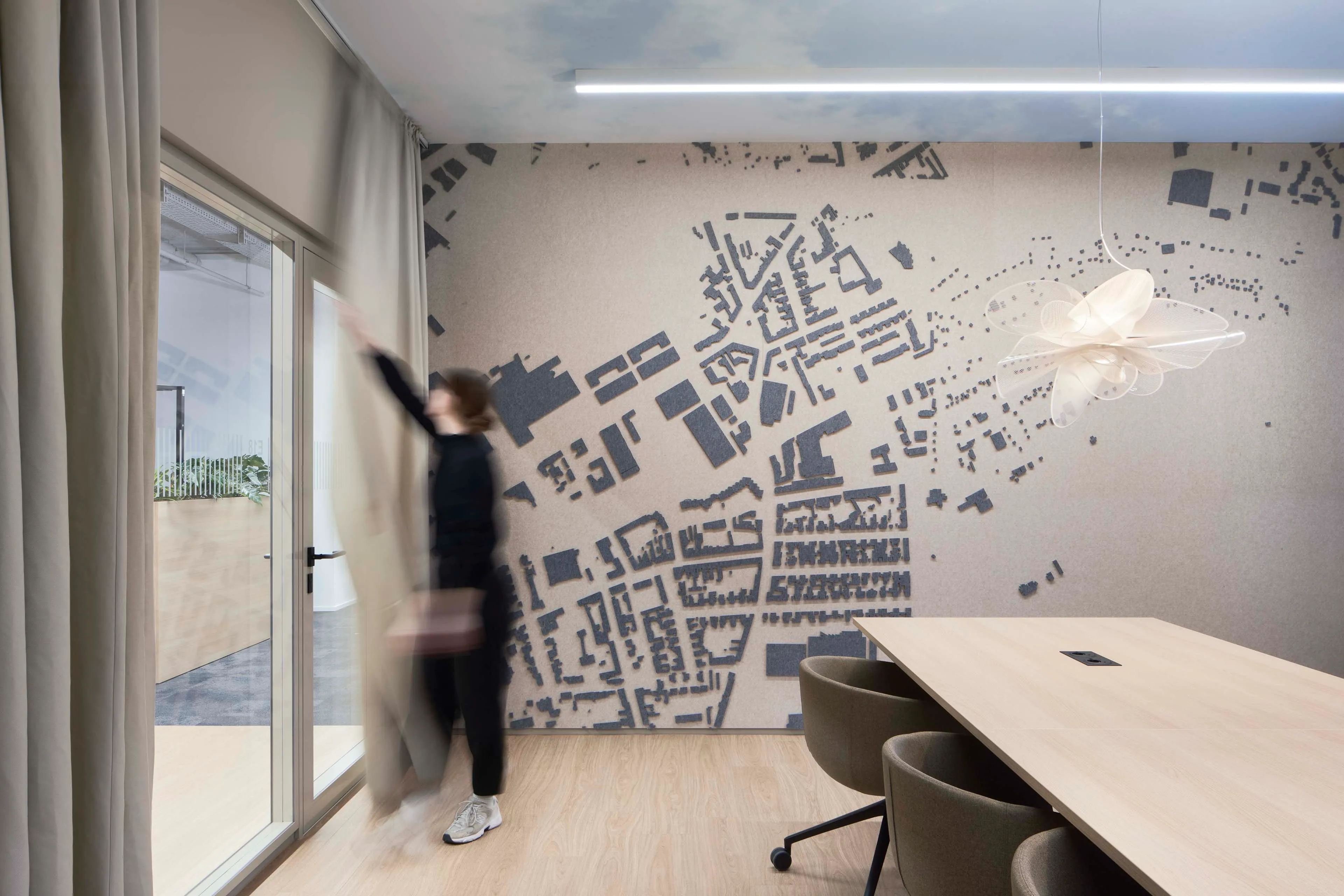 Frau zieht in modernem Bürovortragsraum einen Vorhang vor einer Glastür auf, mit großem Holztisch, Stühlen und Wand mit Stadtplan-Dekoration.