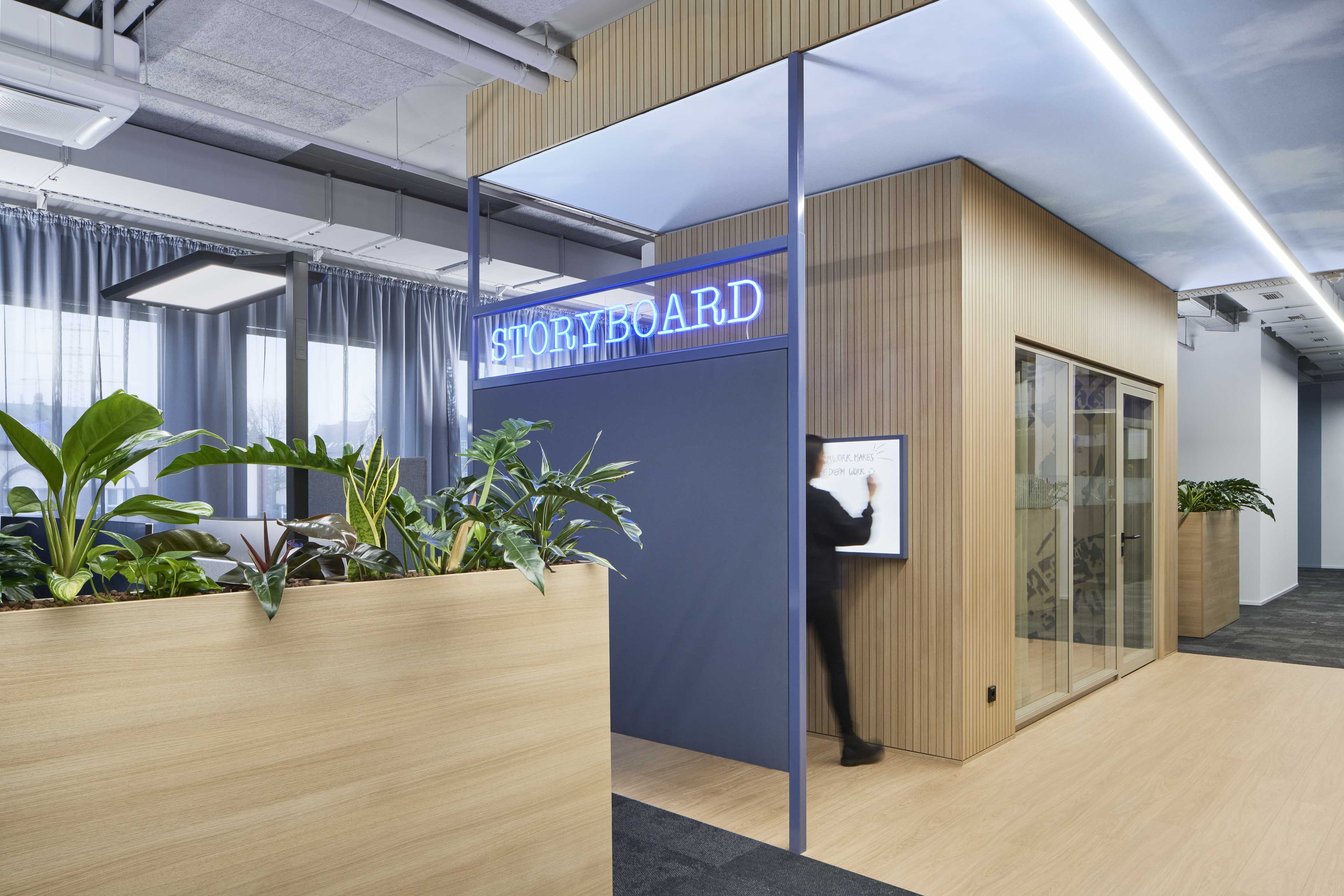 Moderner Büroraum mit Holzverkleidung, Pflanzen und einem Bereich namens Storyboard mit Person, die an einem Whiteboard schreibt.