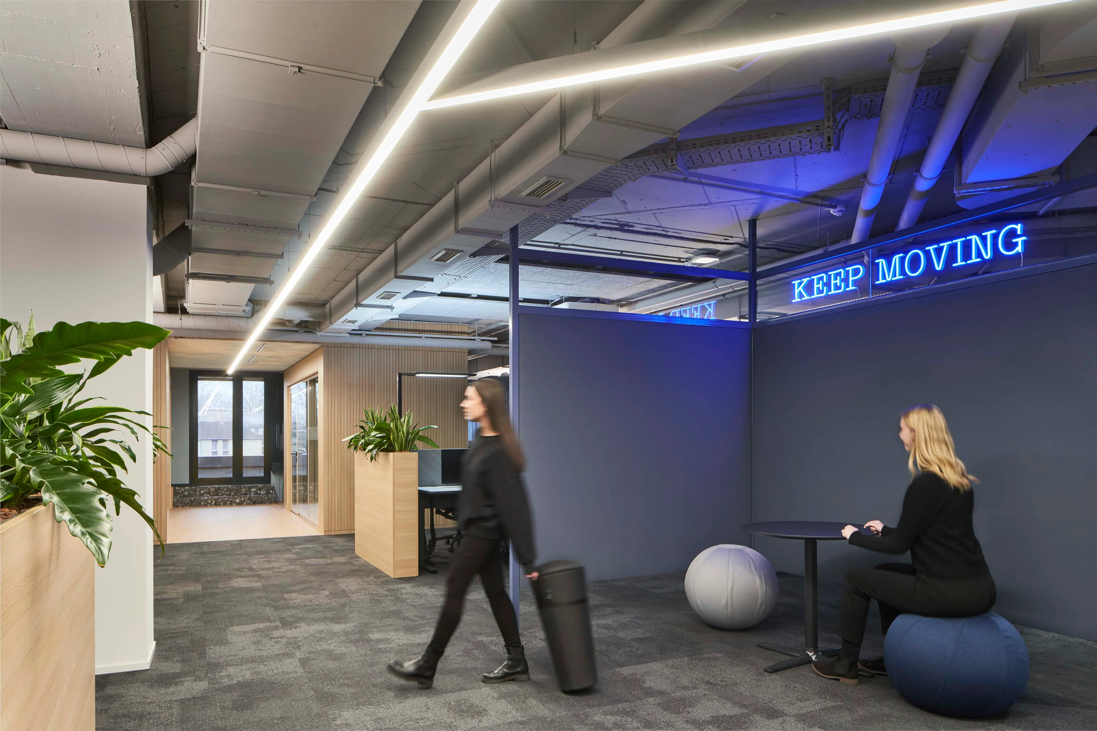 Moderne Bürofläche mit grauem Teppich, zwei Personen, eine sitzt auf einem Sitzball am Tisch, die andere läuft mit einem Koffer, blaues Neon-Schild mit Aufschrift "KEEP MOVING" an der Wand.