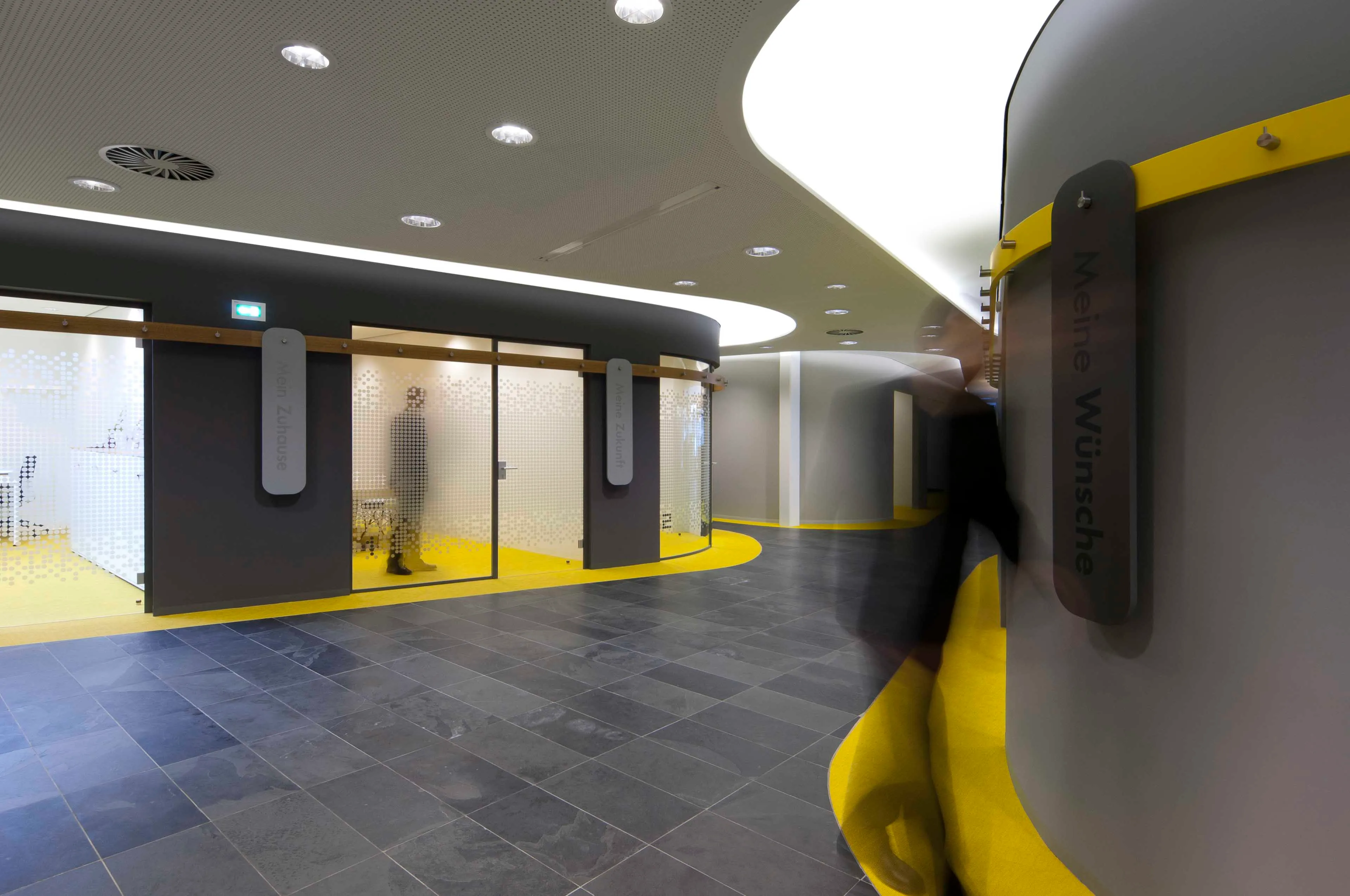 Moderne Innenraumgestaltung mit geschwungenem Flur, gelbem Bodenbelag, grauen Wänden und Glasbüros, Person in Bewegung unscharf.