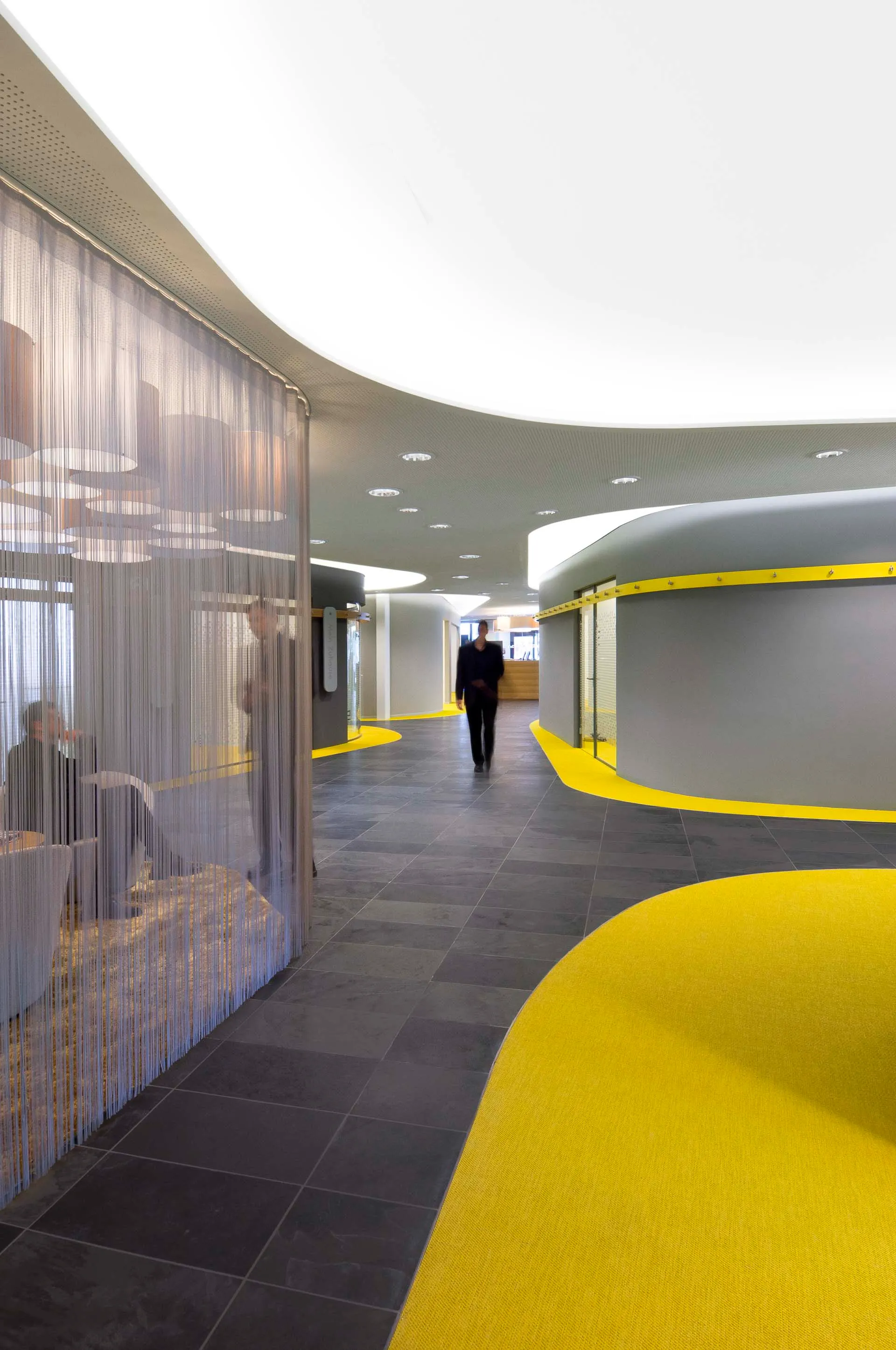 Moderner Innenraum mit gewelltem gelben Teppich, dunklem Fliesenboden, abgerundeten grauen Wänden und einer abgehängten Decke mit runden Lichtquellen, eine Person geht im Gang, hinter einer durchsichtigen Vorhangwand sitzen zwei Personen.