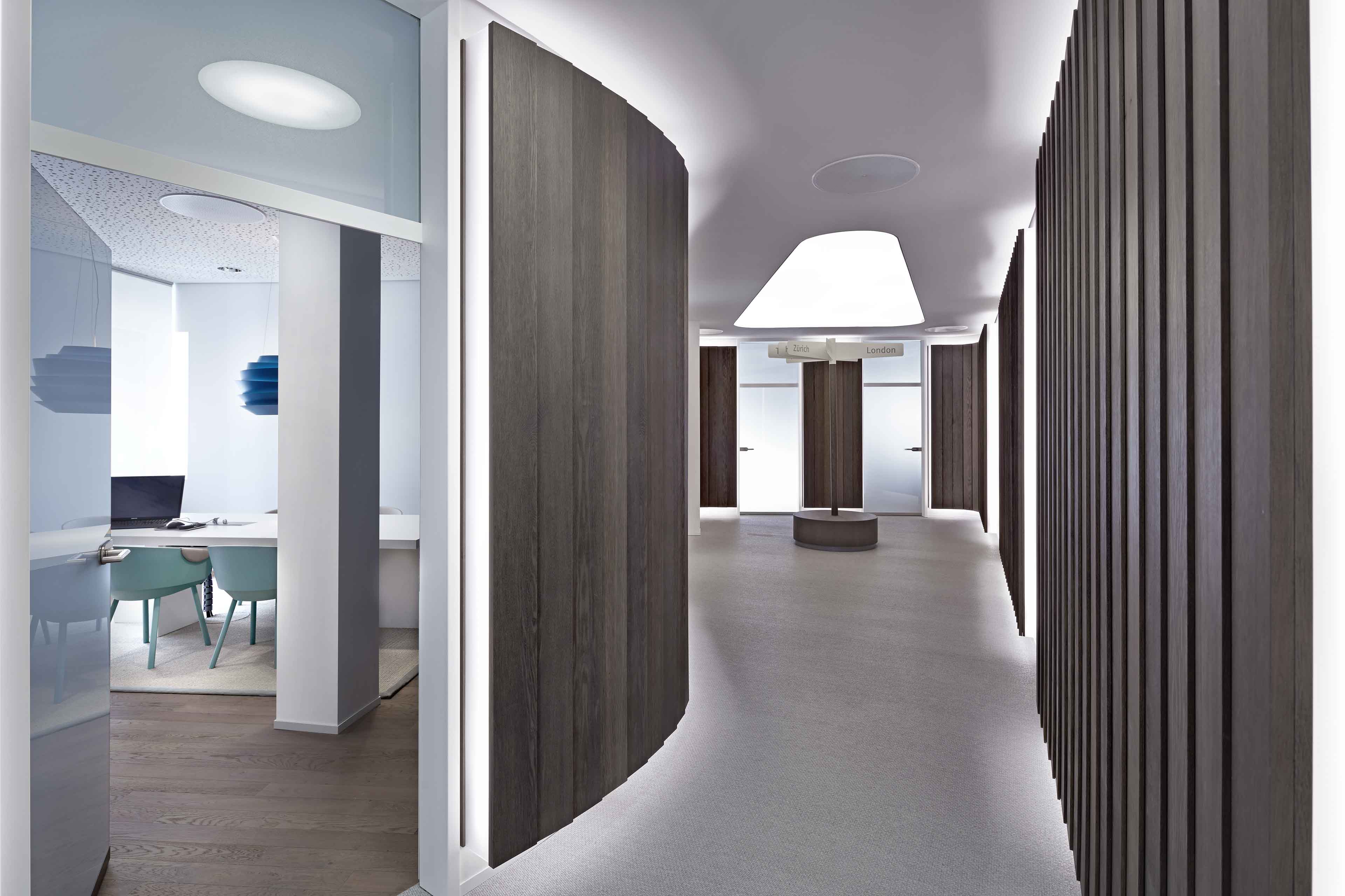 Moderner Büroflur mit geschwungenen Holzwänden, heller Deckenleuchte und beschriftetem Wegweiser zu Zürich und London.