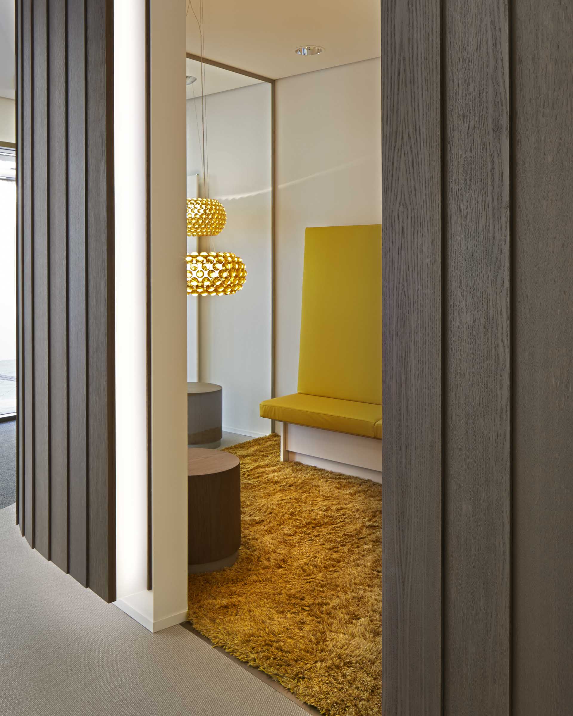 Moderner Sitzbereich mit gelbem gepolstertem Sitz, zwei runden Holztischen, orangem Teppich und zwei goldfarbenen Hängeleuchten.
