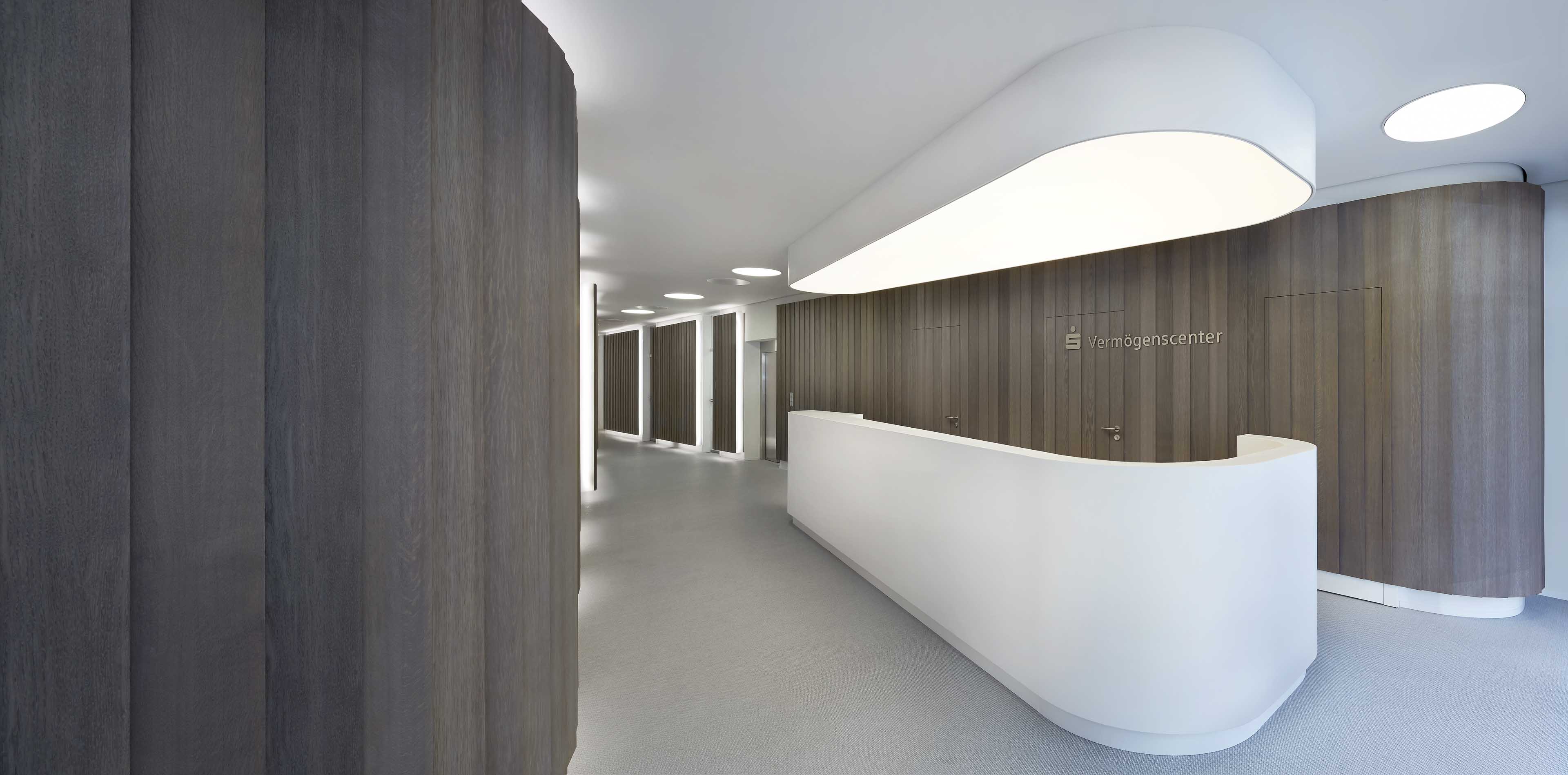 Modernes Büro mit weißem Empfangstresen und dunklen Holzpaneelen, Schild mit der Aufschrift ‚Vermögenscenter‘ an der Wand.