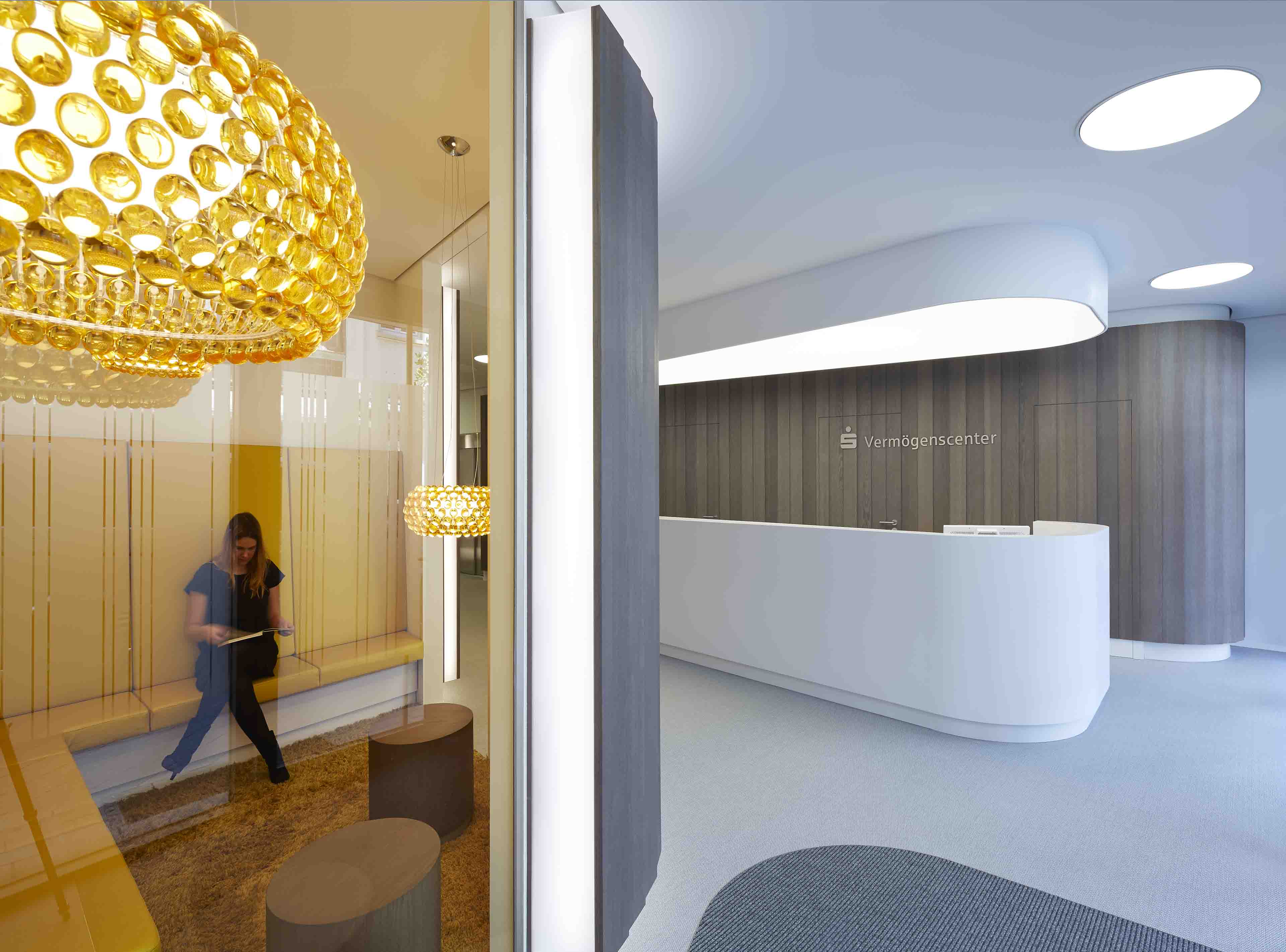 Moderne Empfangstheke mit geschwungenem weißen Tresen und Holzverkleidung, davor eine Glaswand mit einem beleuchteten Sitzbereich, in dem eine Frau sitzt und liest.