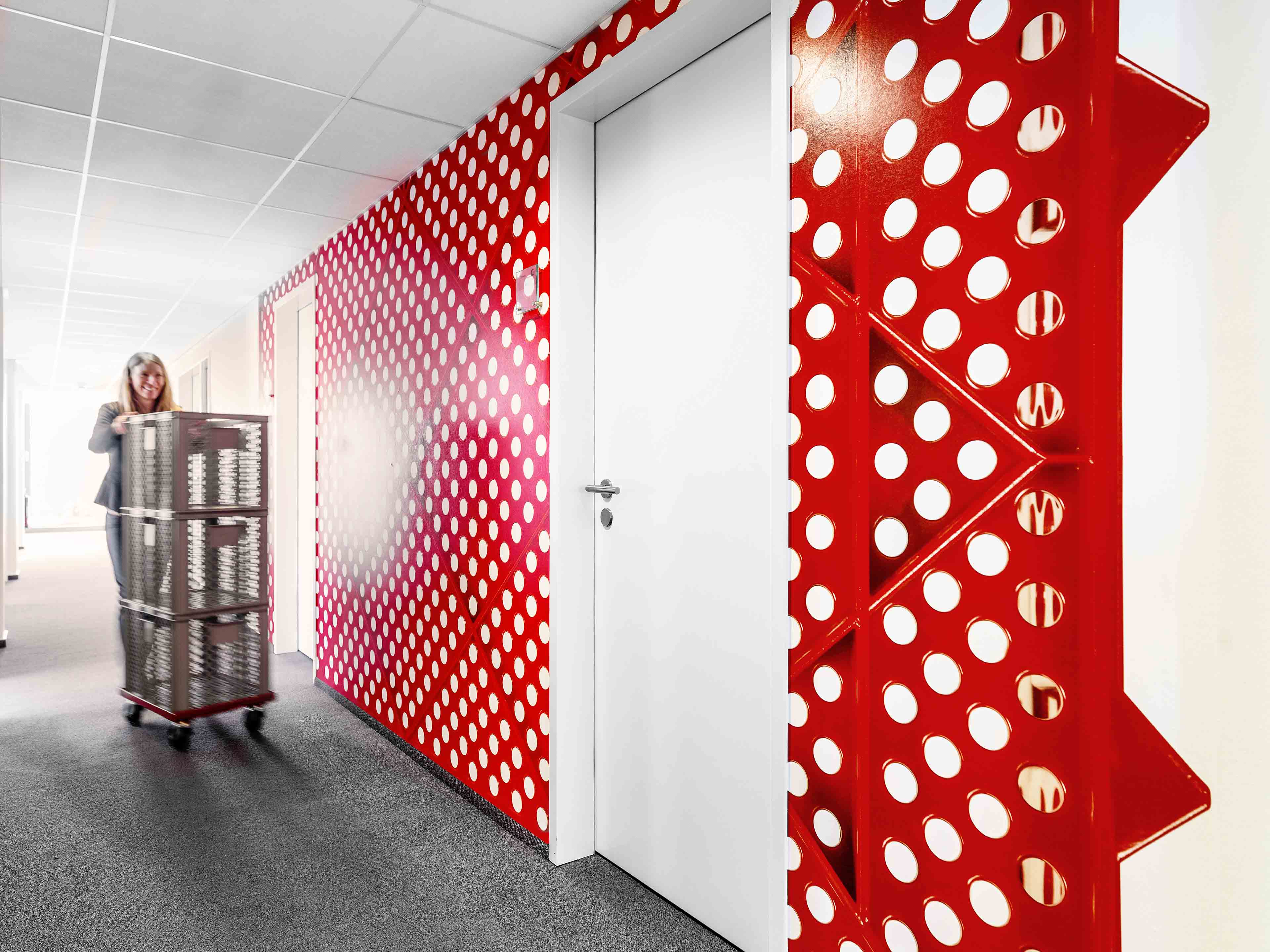 Frau schiebt einen Wagen mit Kisten durch einen Büroflur mit einer roten Wand mit weißen Punkten.