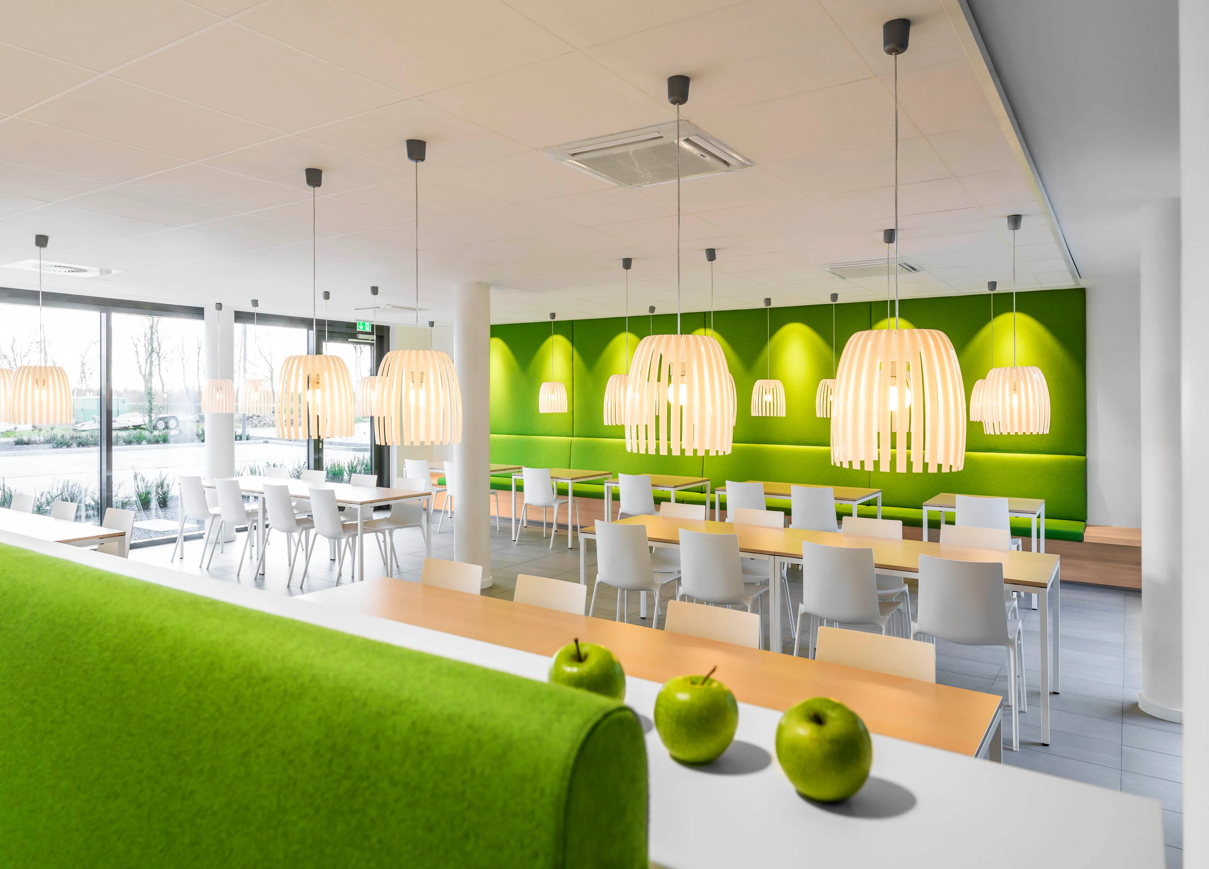Moderne Cafeteria mit weißen Stühlen, holzfarbenen Tischen, grünen Wandpolstern, hängenden Lampen und drei grünen Äpfeln im Vordergrund.