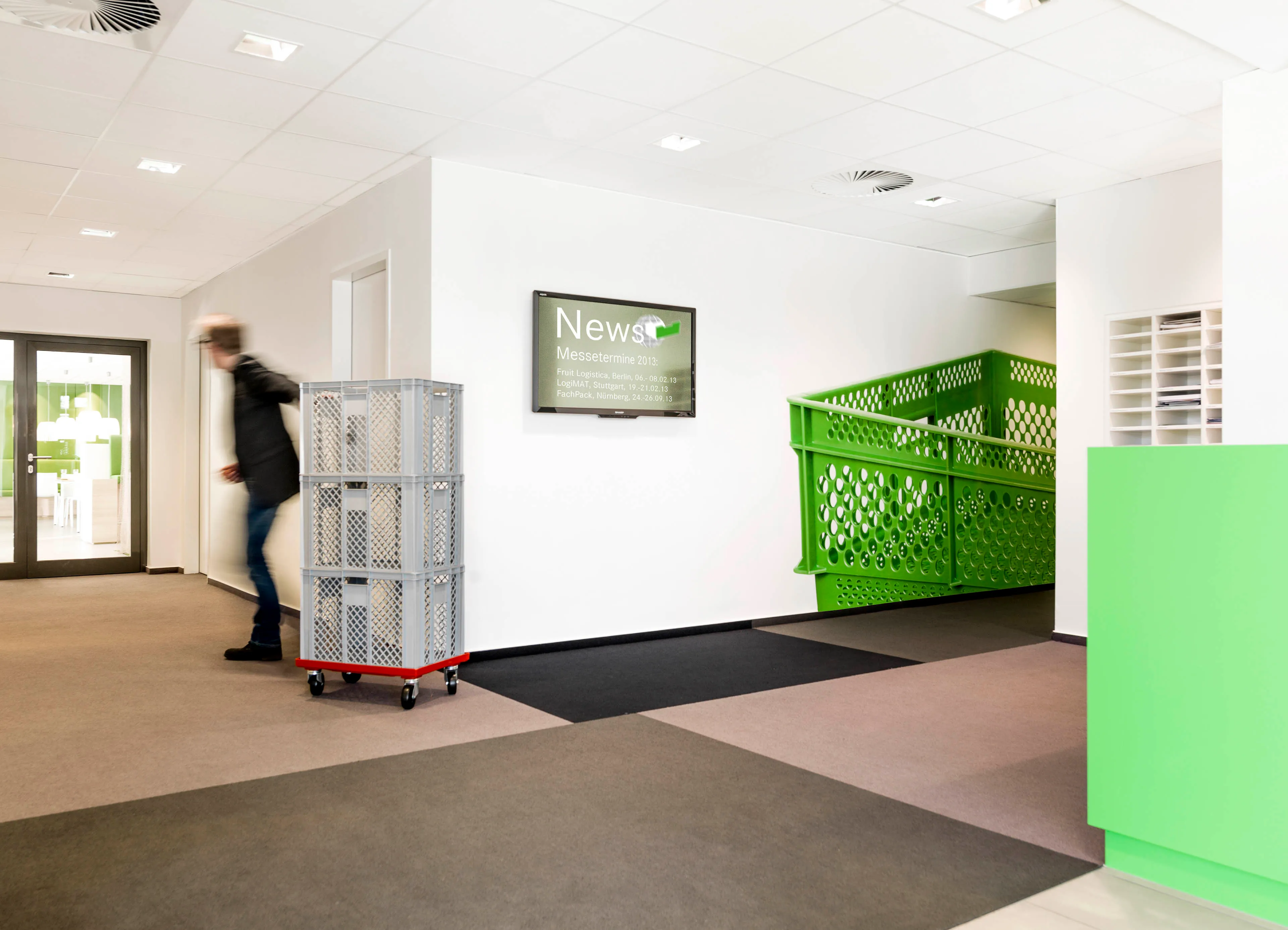 Büro mit grünem Treppengeländer, einem Mann in Bewegung und gestapelten grauen Kisten auf einem roten Rollwagen.