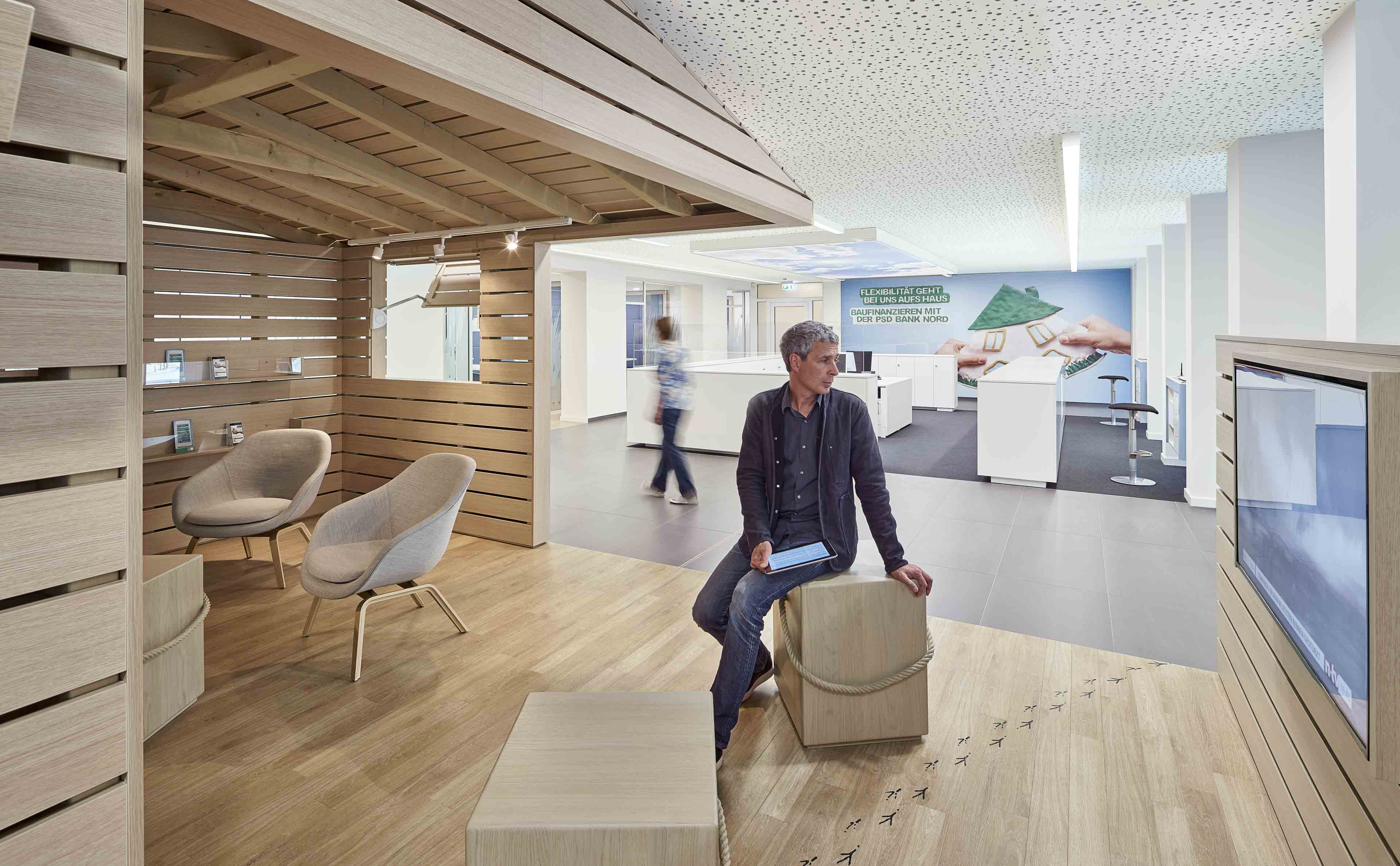 Moderner Wartebereich in einer Bank mit Holzverkleidungen, Sitzwürfeln, zwei Sesseln und einem Mann, der auf einem Würfel sitzt und ein Tablet hält.