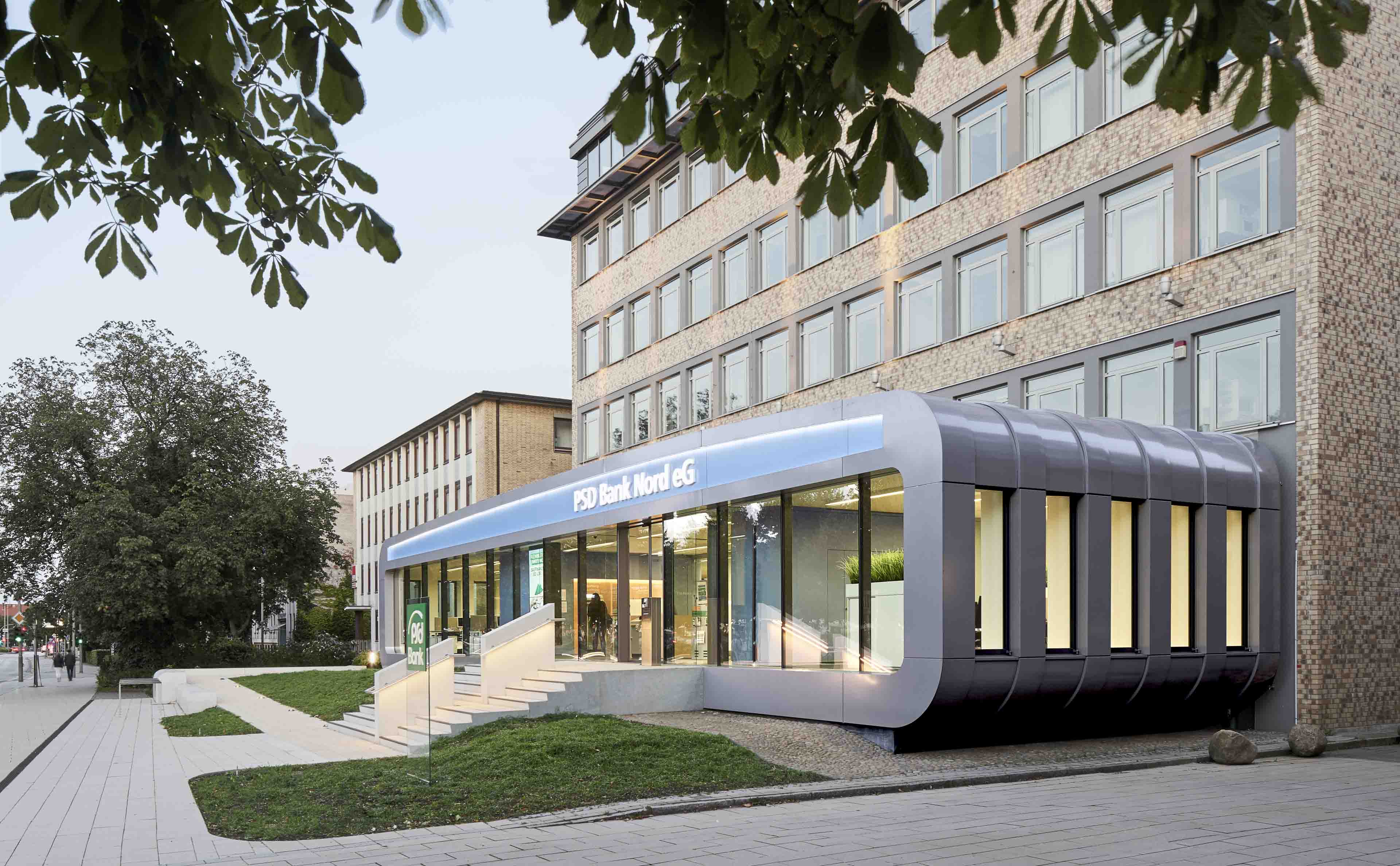 Moderner Eingang der PSD Bank Nord eG mit beleuchteten Glasfenstern und Treppe vor einem mehrstöckigen Backsteingebäude.