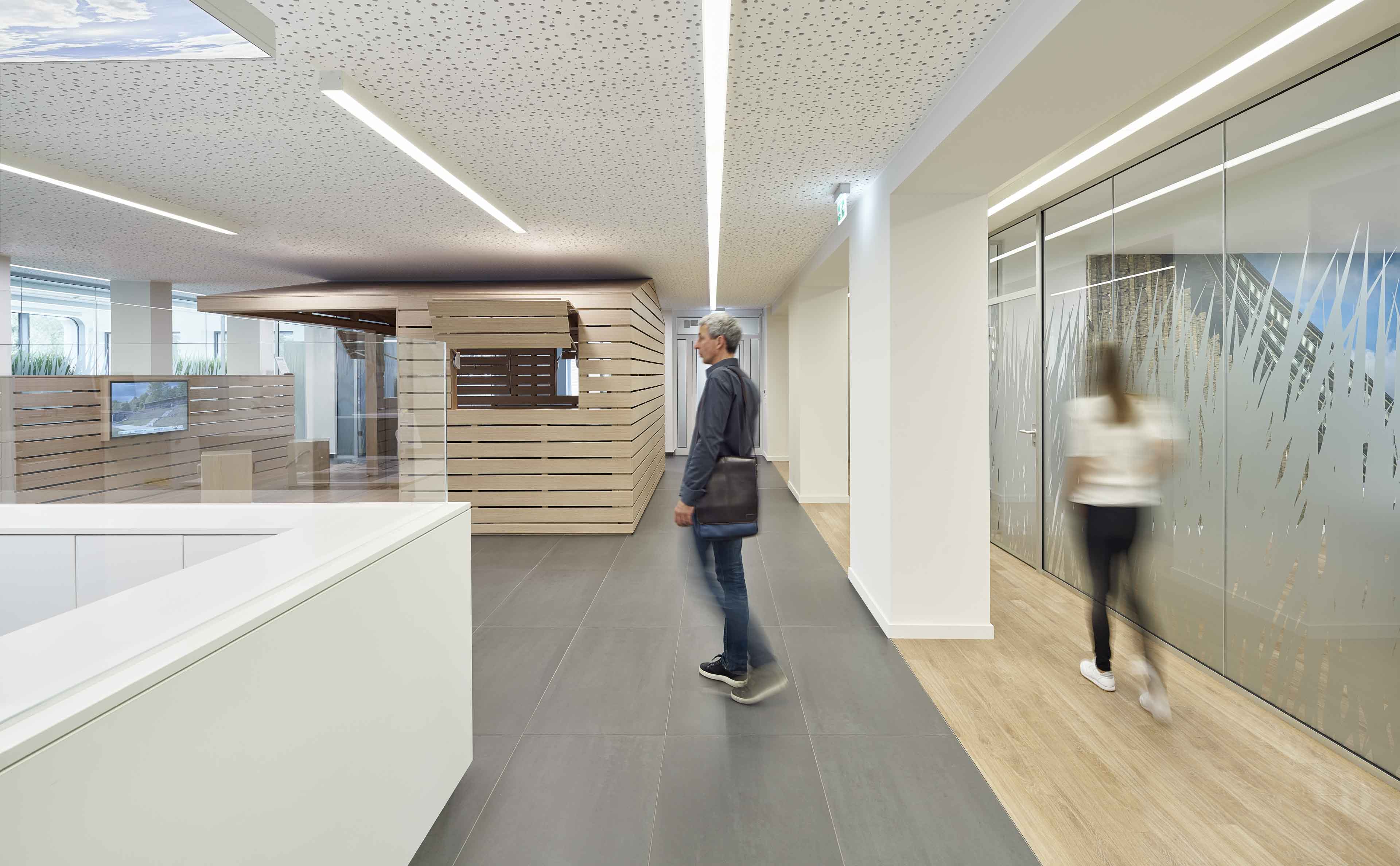 Moderner Büroraum mit grauem Boden, Holzpaneelen, Glaswänden mit Muster und zwei unscharfen Personen in Bewegung.
