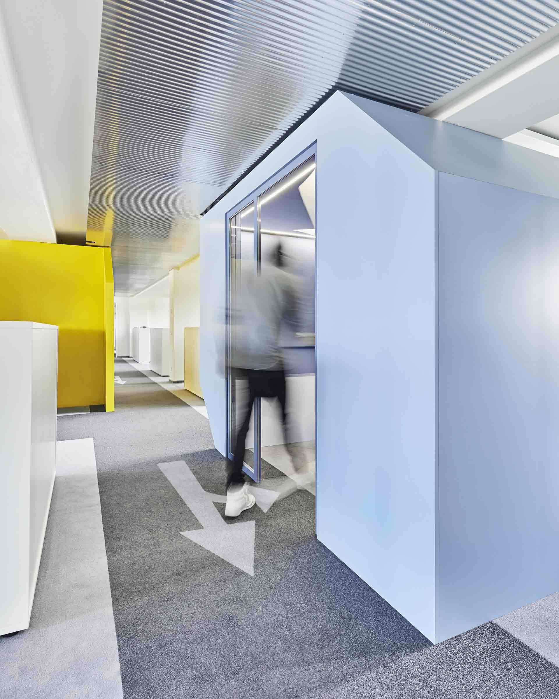 Verschwommene Person betritt einen hellblauen Meetingraum in einem modernen Büro mit gelben Wänden und grauem Teppichboden.
