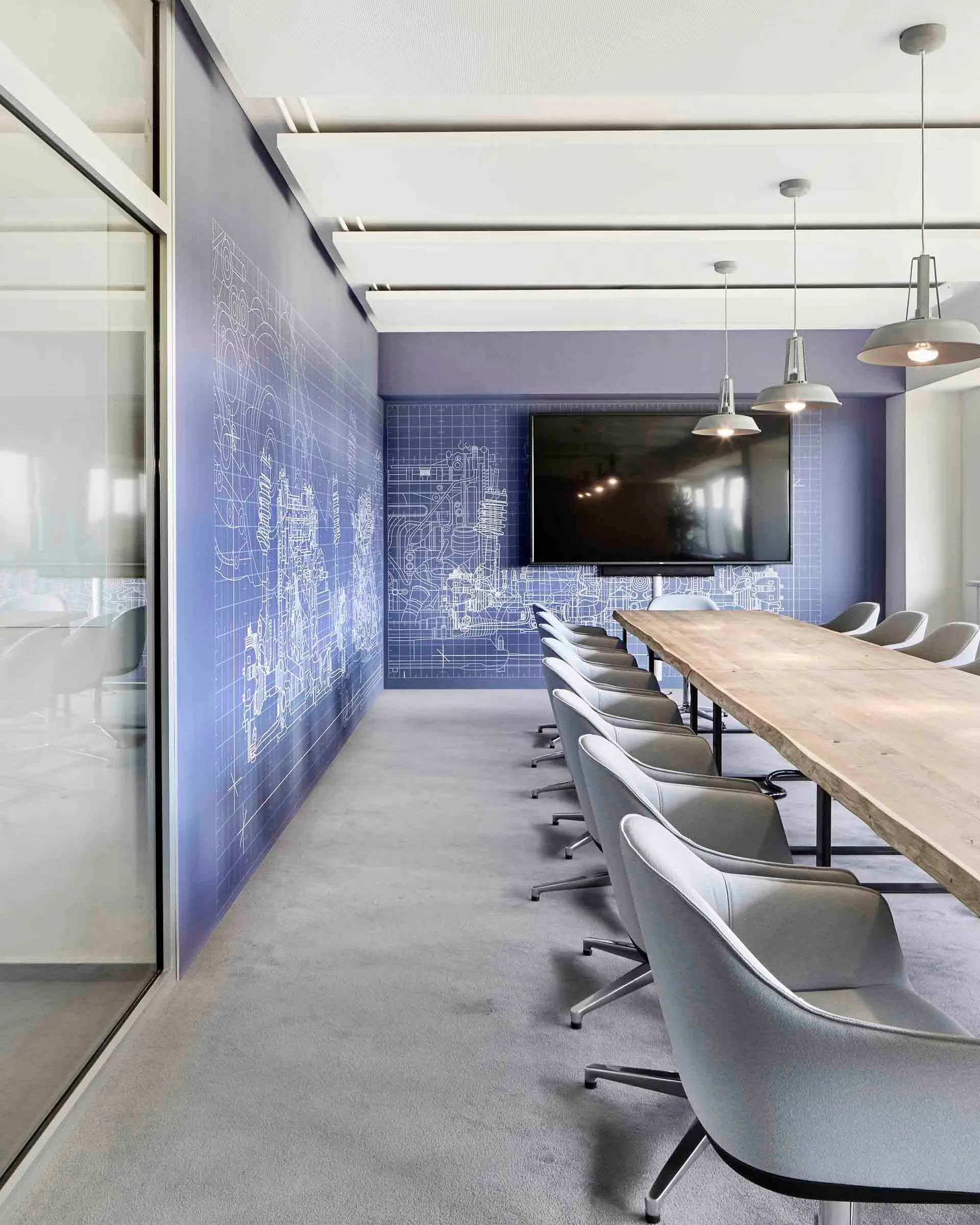 Moderner Konferenzraum mit langem Holztisch, grauen Stühlen, einem großen Bildschirm an der Wand und technischen Zeichnungen auf blauer Wand.