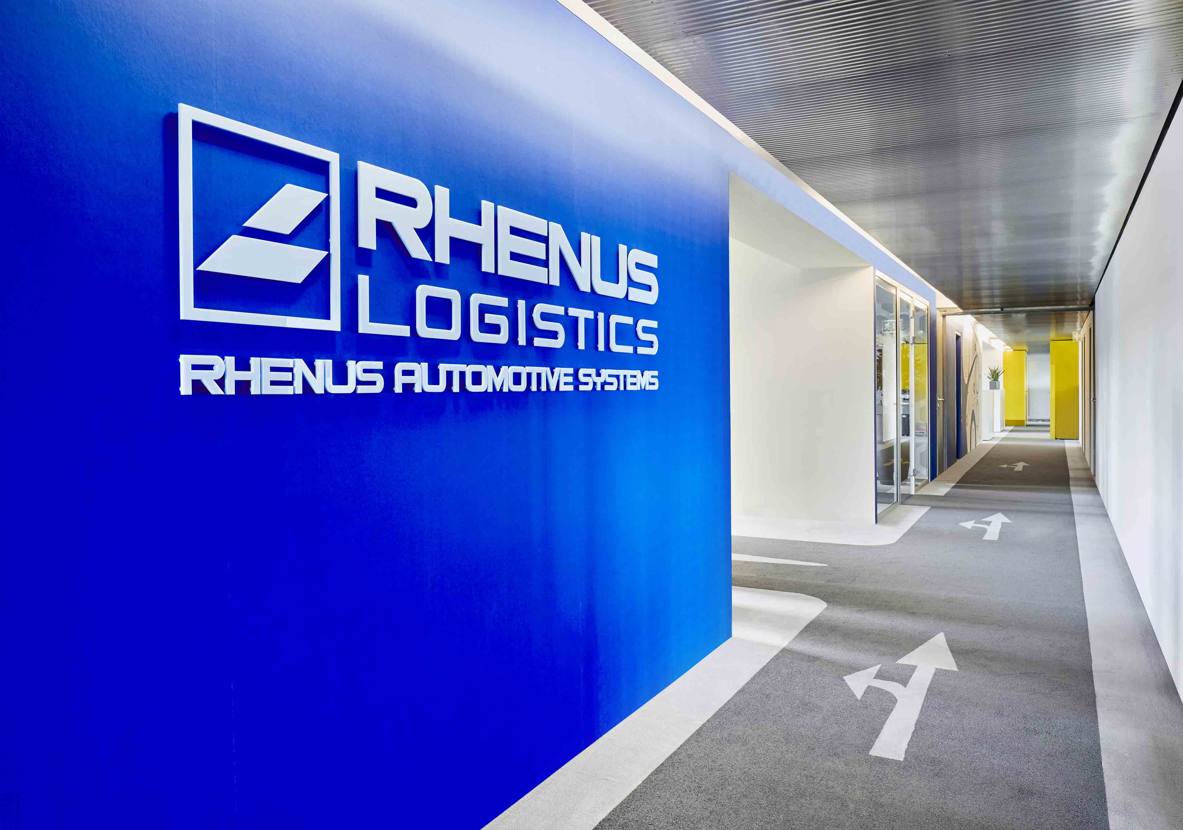Flur mit blauem Logo von Rhenus Logistics an der linken Wand und Pfeilen auf dem Boden, der zu gelben Türen am Ende des Gangs führt.