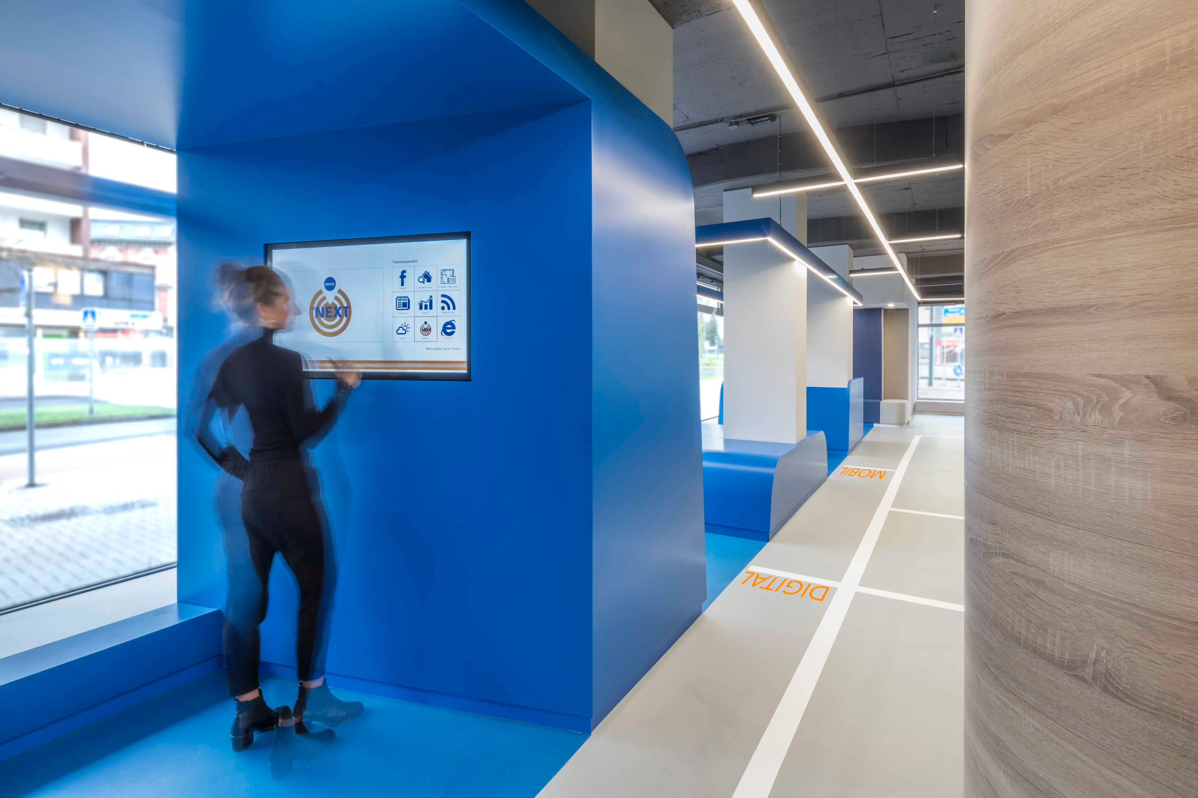 Frau bedient einen Touchscreen in einem modernen, hellen Raum mit blauen und weißen Elementen.