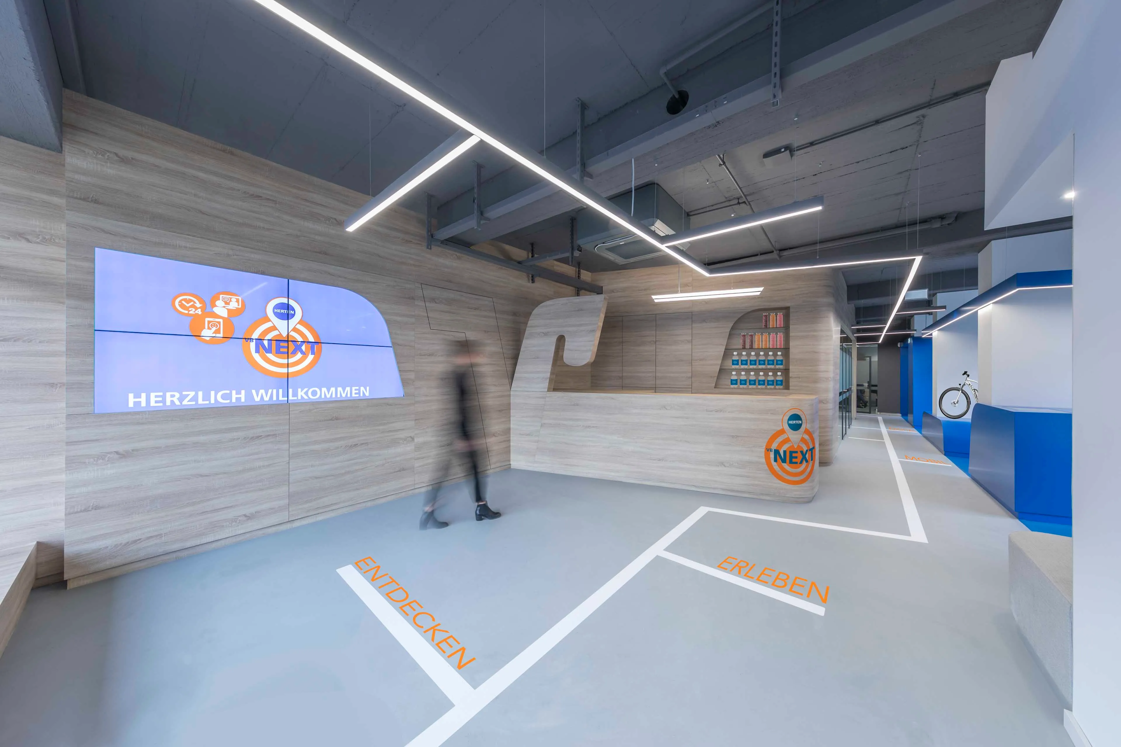 Moderner Empfangsbereich mit Holzverkleidung und digitaler Anzeige, auf dem Boden stehen die Worte 'ENTDECKEN' und 'ERLEBEN'.