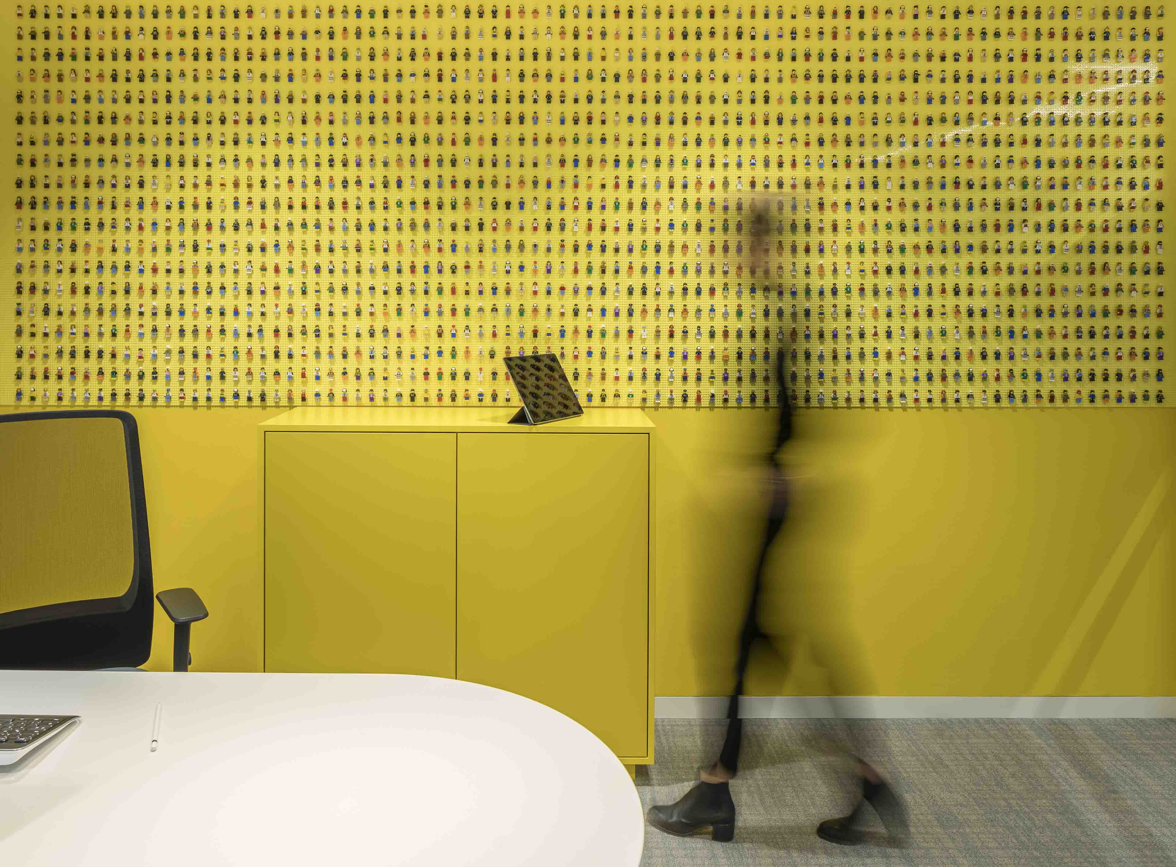 In einem gelben Büro steht eine gelbe Kommode vor einer Wand mit vielen kleinen LEGO-Figuren, während eine verschwommene Person vorbeigeht.