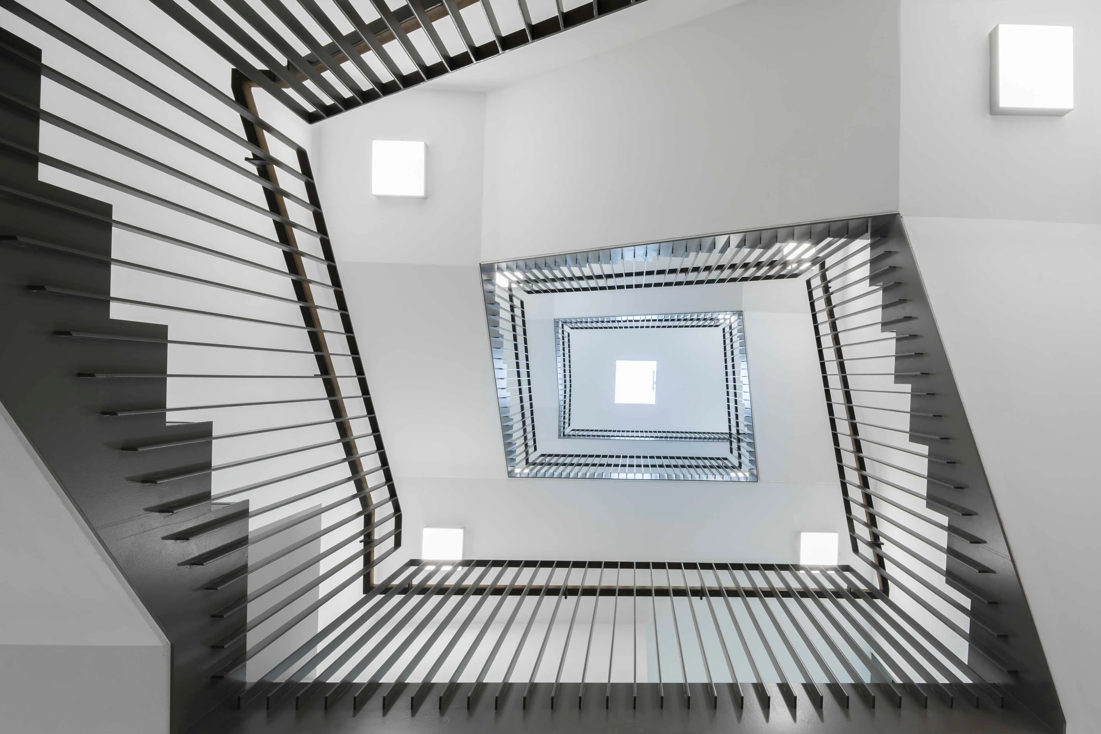 Blick nach oben durch ein modernes, quadratisches Treppenhaus mit schwarzen Geländern und weißen Wänden.