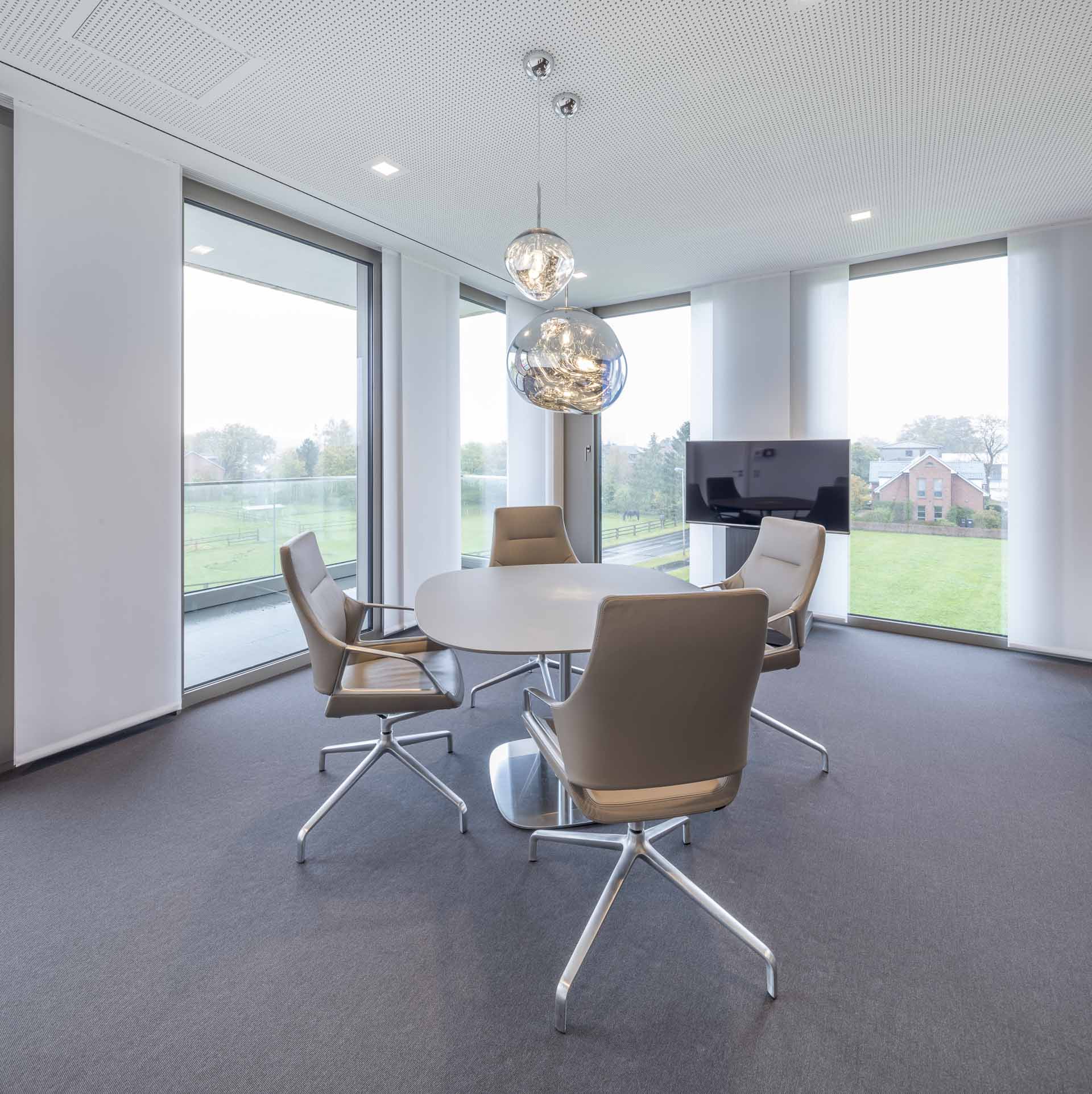 Moderner Besprechungsraum mit rundem Tisch, vier drehbaren Stühlen und großen Fenstern mit Blick auf eine grüne Landschaft.