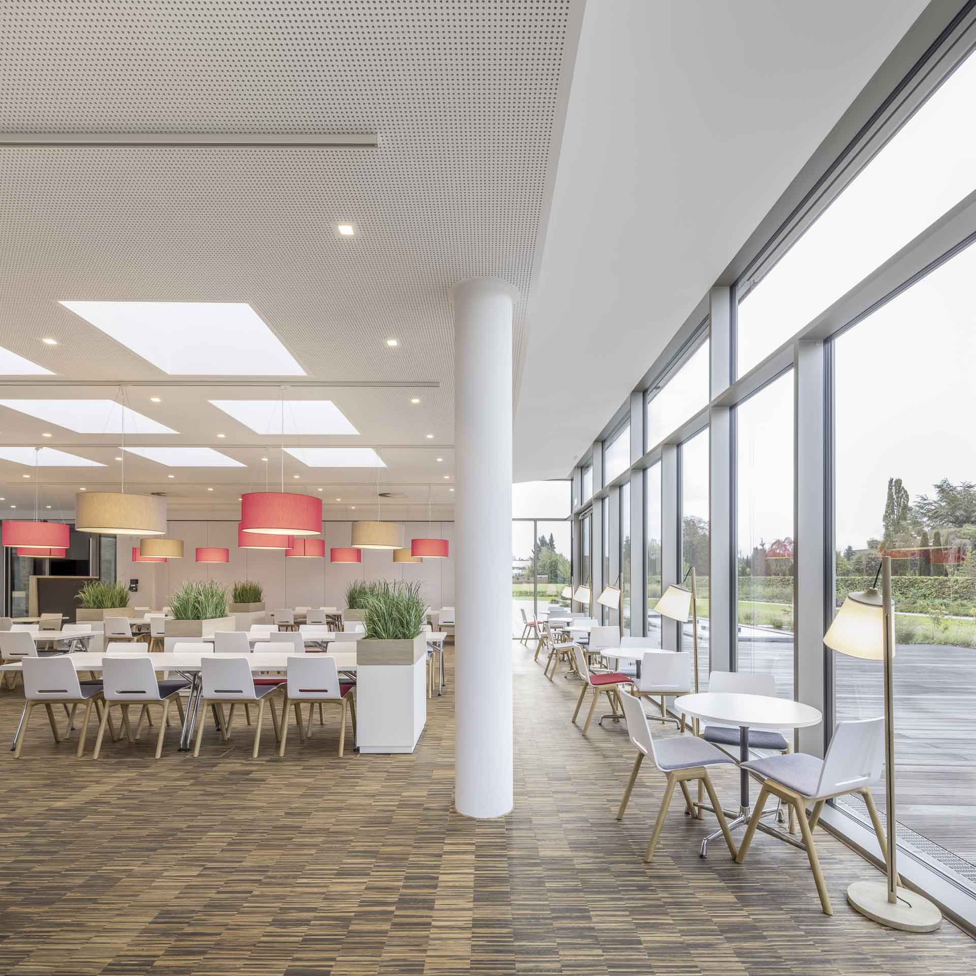 Moderner Speiseraum mit großen Fenstern, weißen Tischen, Stühlen mit Holzbeinen und roten sowie beigen Hängelampen.