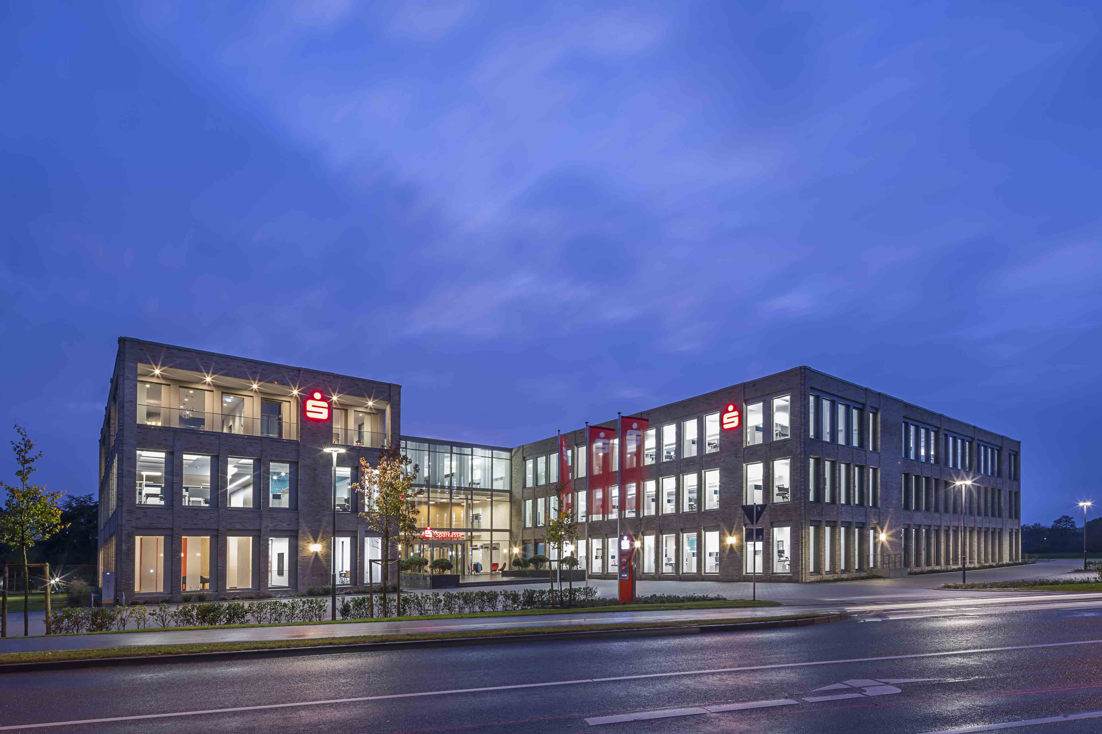 Beleuchtetes modernes Bankgebäude der Sparkasse bei Dämmerung mit großen Fenstern und roten Logos.