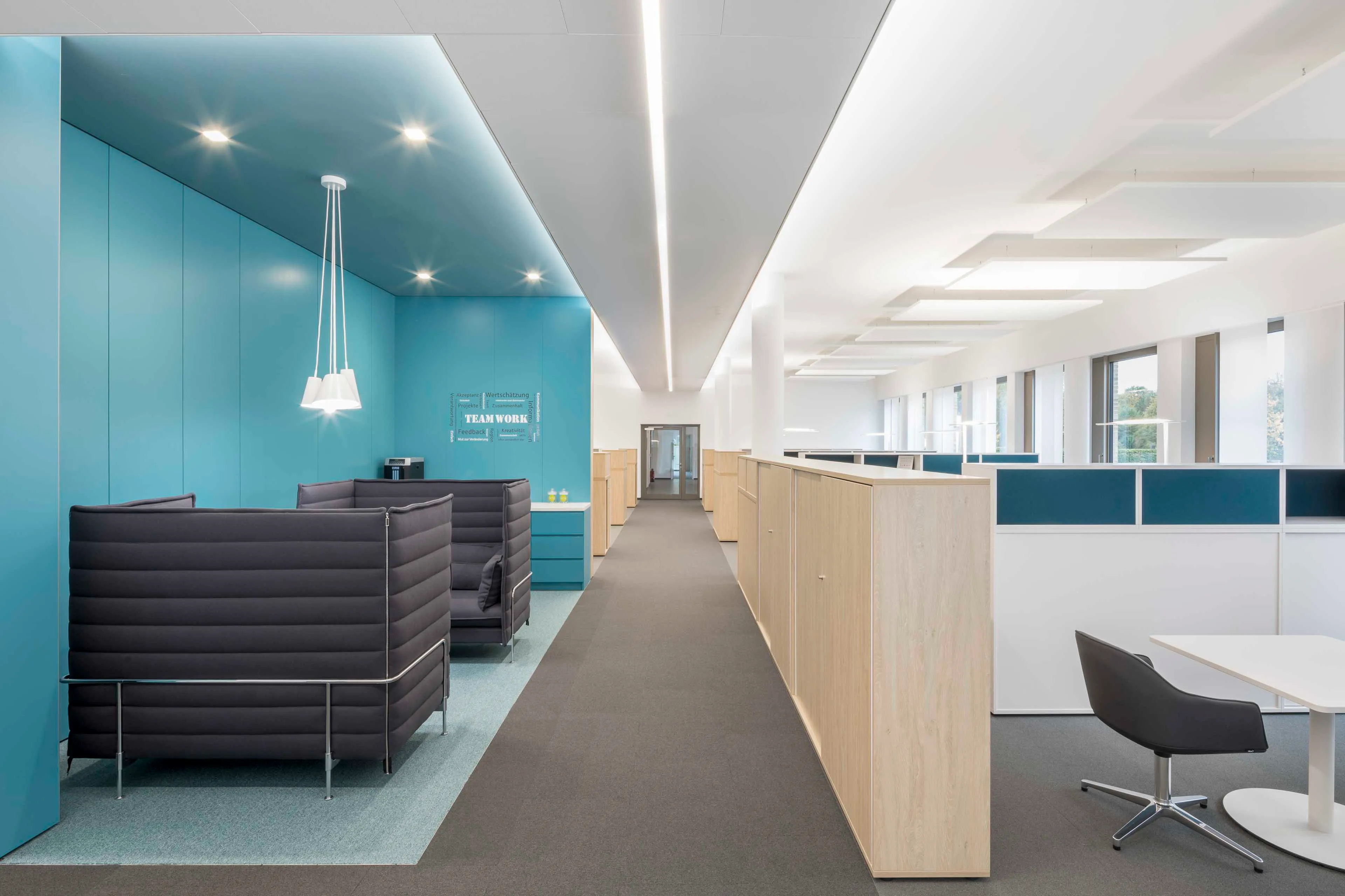 Moderner Büroraum mit blauen Trennwänden, grauen Sitznischen, Holzmöbeln und einer langen Beleuchtung an der Decke.