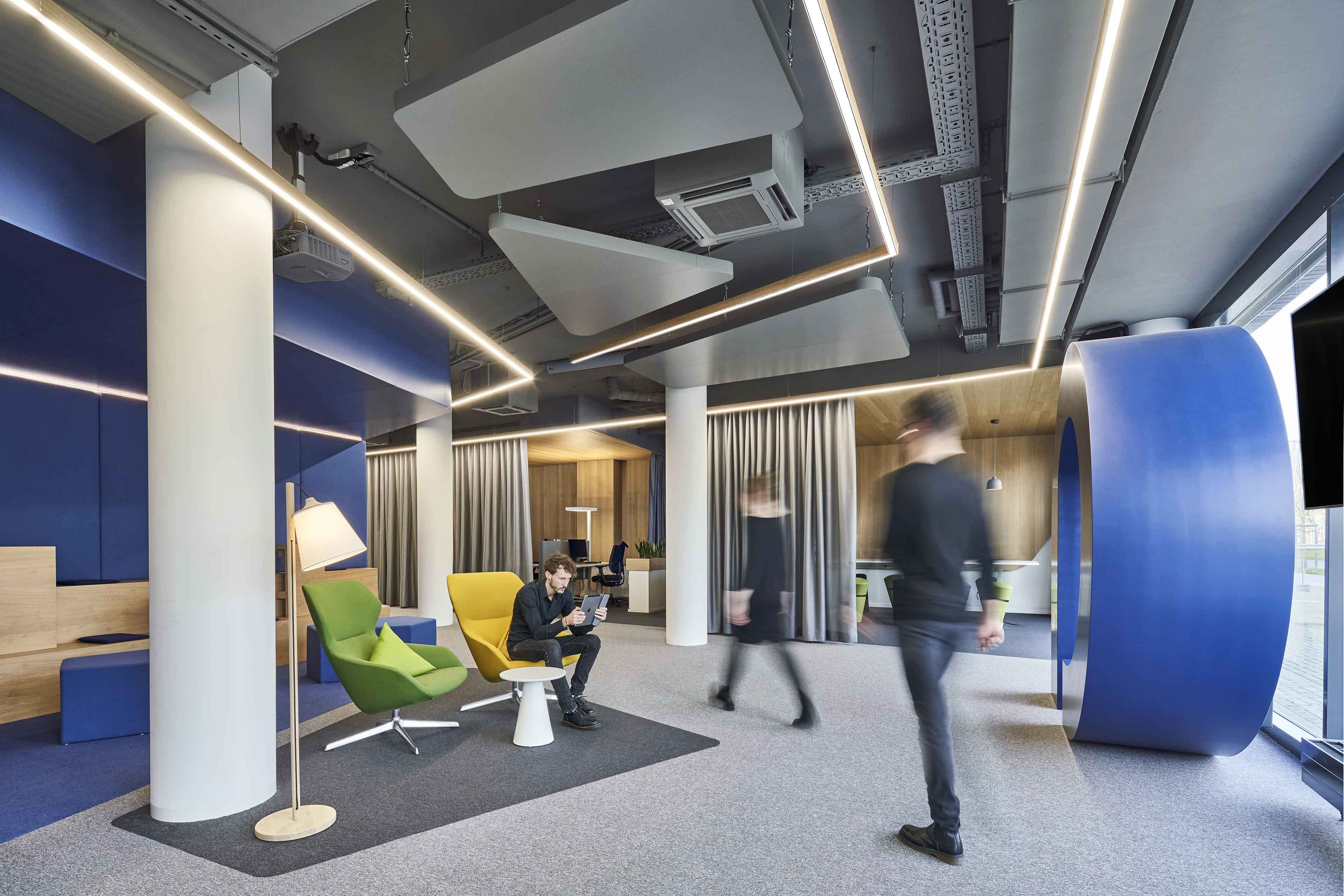 Modernes Büro mit bunten Sesseln, beleuchtetem Stehlampelement, und zwei unscharfen Personen, die vorbeigehen.