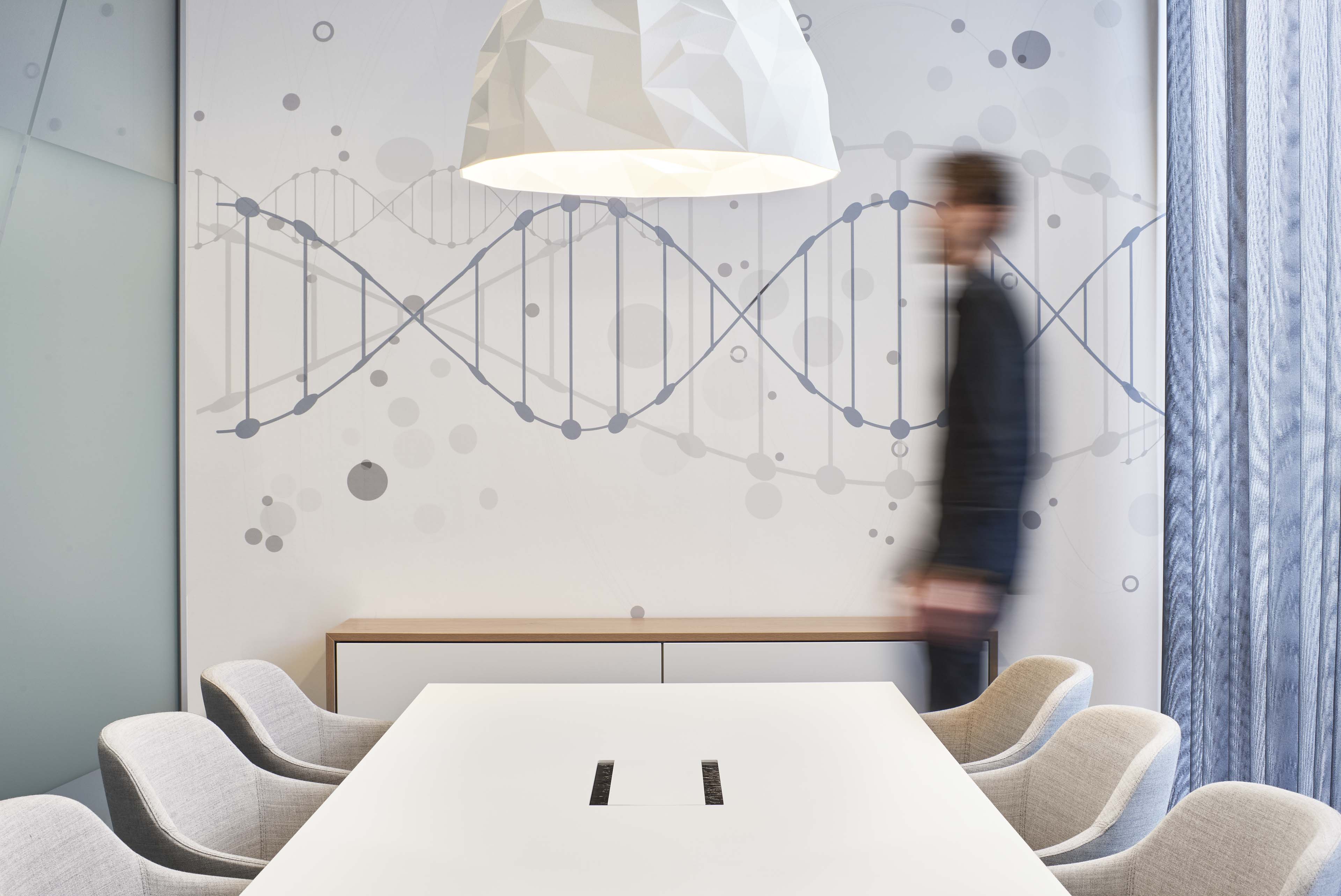 Moderner Konferenzraum mit weißem Tisch, grauen Stühlen, DNA-Doppelhelix-Wandbild und verschwommenem Mann im Hintergrund.