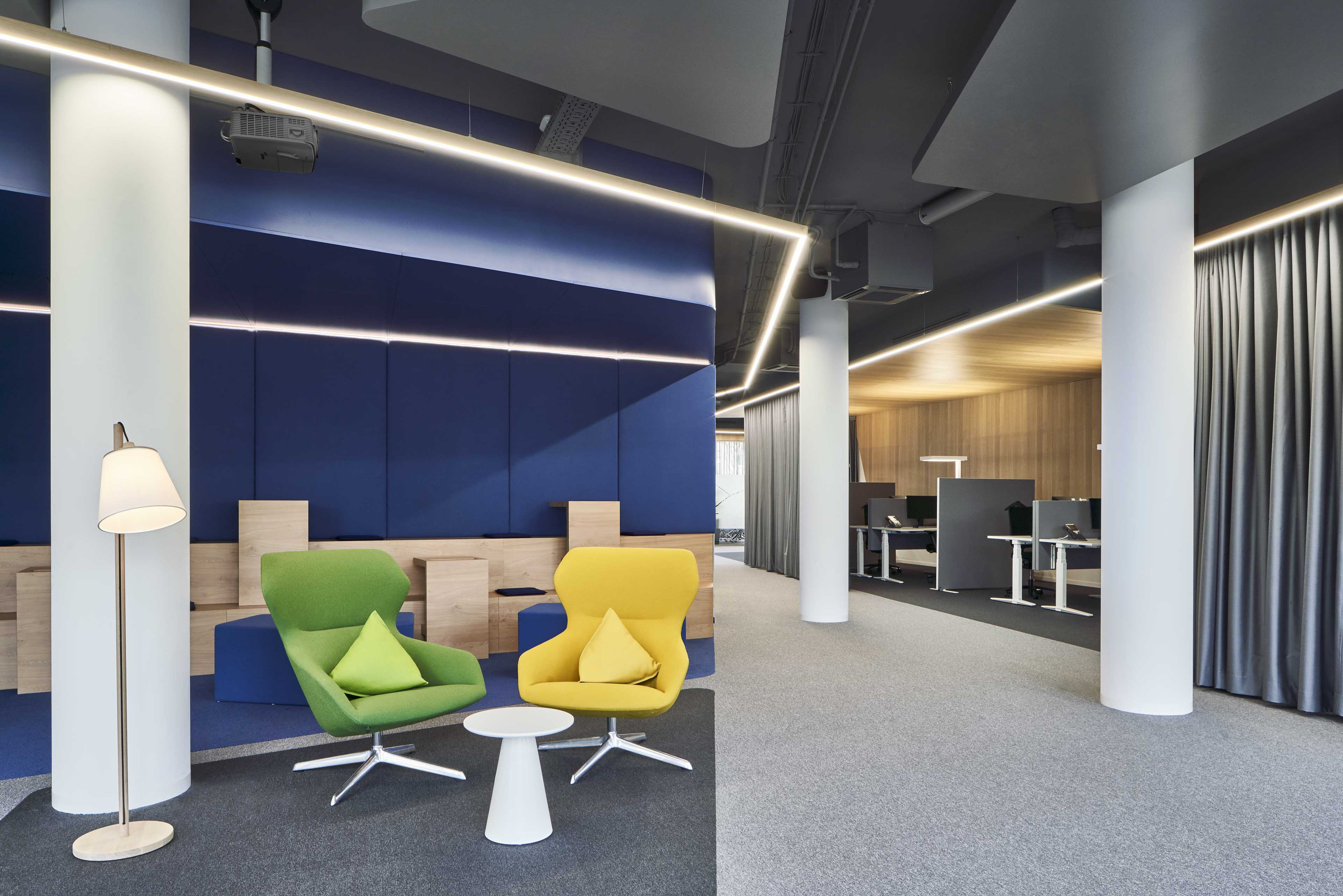 Modernes Büro mit grünen und gelben Sesseln, einem weißen Tisch, blauen Wandpaneelen und Arbeitsplätzen im Hintergrund.