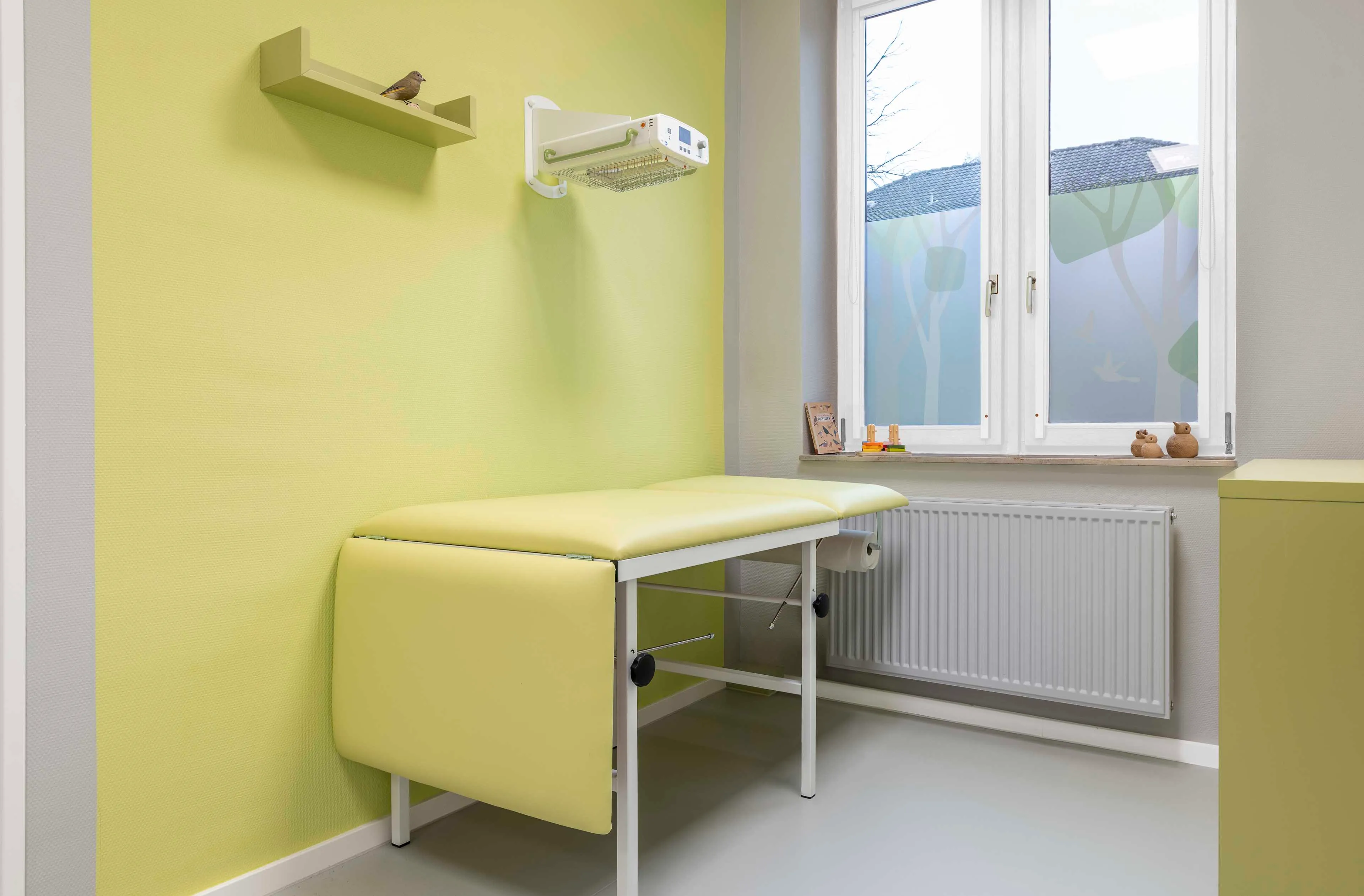Helles Behandlungszimmer mit gelber Liege, kleinem Vogel auf einem Wandregal und Fenster mit Sichtschutzfolie.