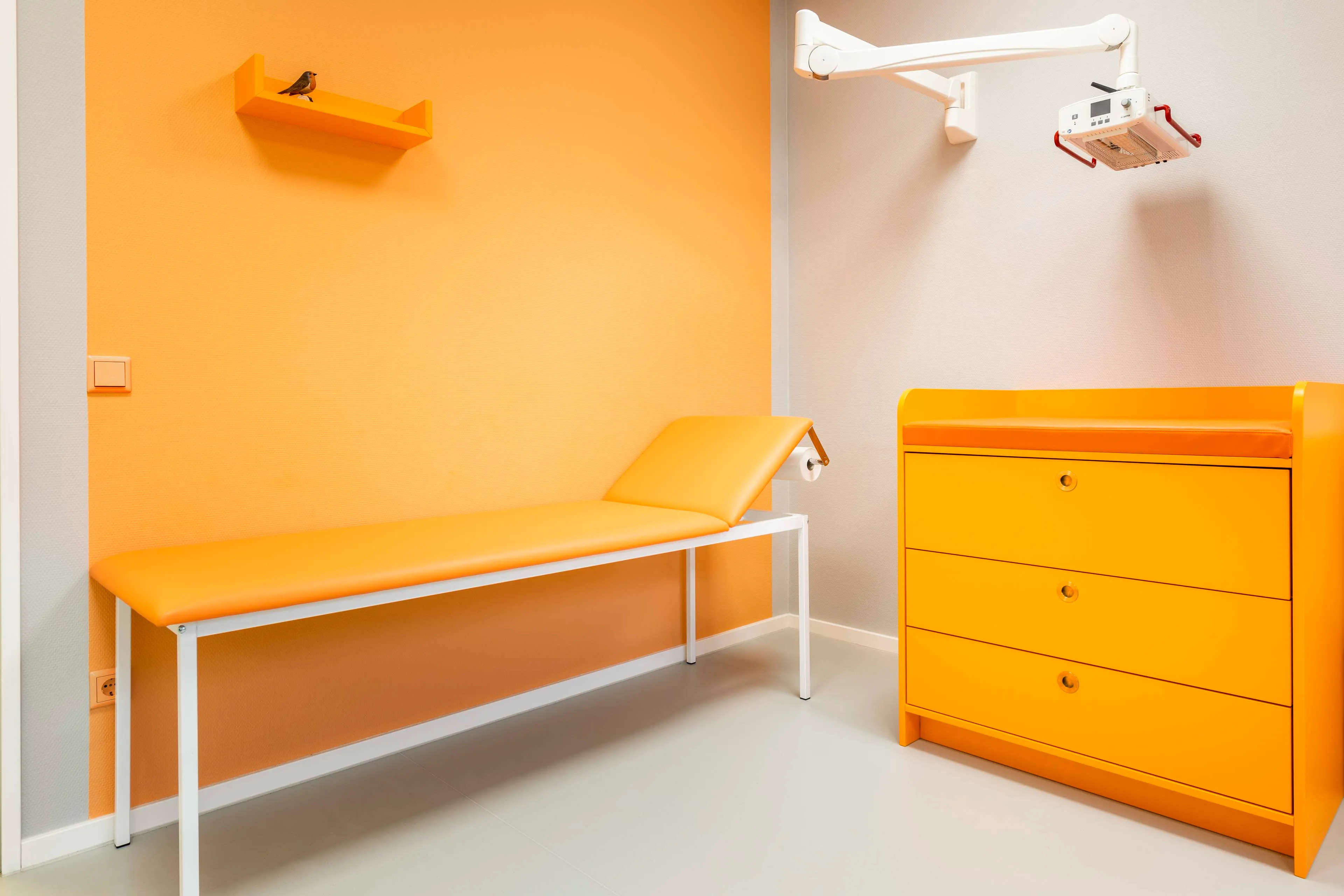Helle Untersuchungsliege mit orangenem Polster vor einer orangefarbenen Wand und gelber Kommode in einem medizinischen Behandlungsraum.