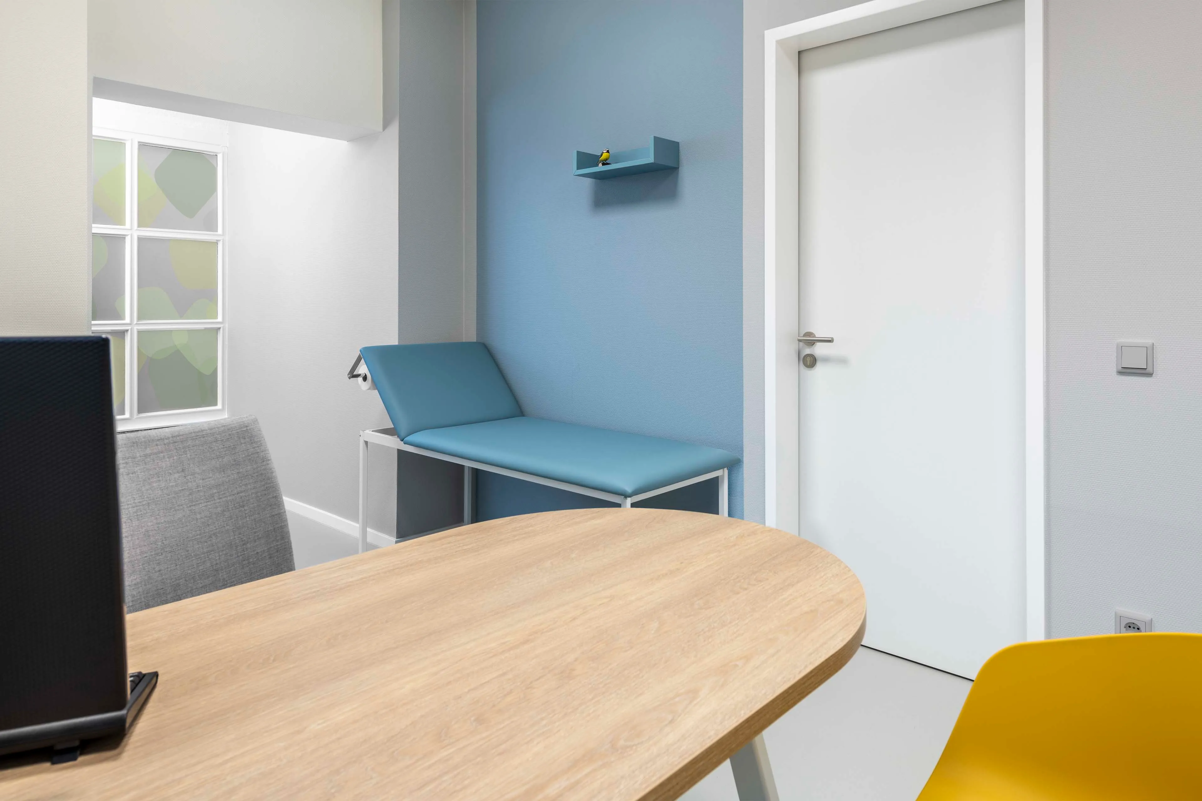 Helles, modernes Untersuchungszimmer mit blauem Liegepolster, Holztisch und gelbem Stuhl.