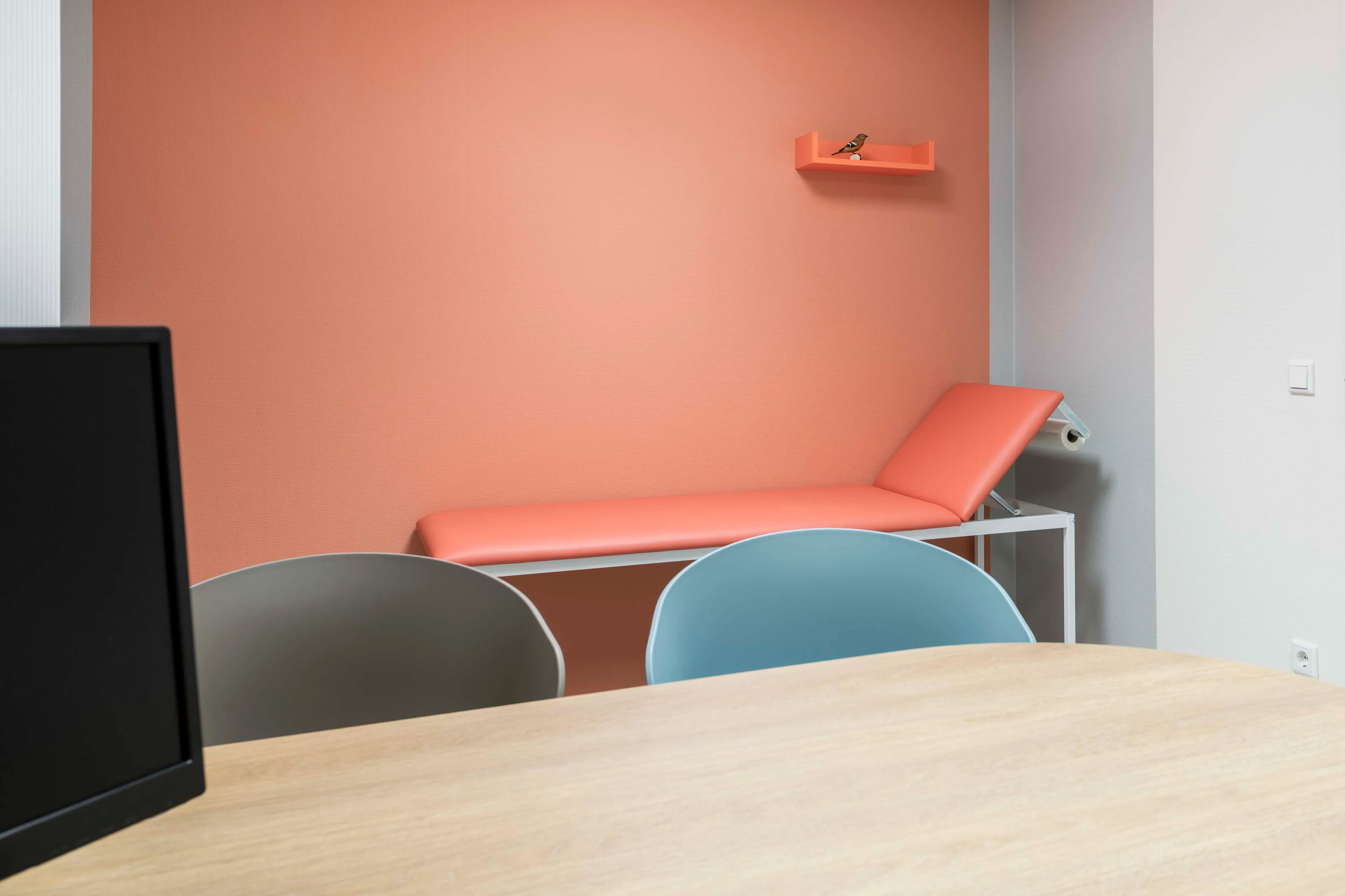Behandlungszimmer mit einem orangefarbenen Untersuchungsliege, zwei Stühlen in Grau und Blau, einem Holztisch und orangefarbenem Wandregal mit Vogel.