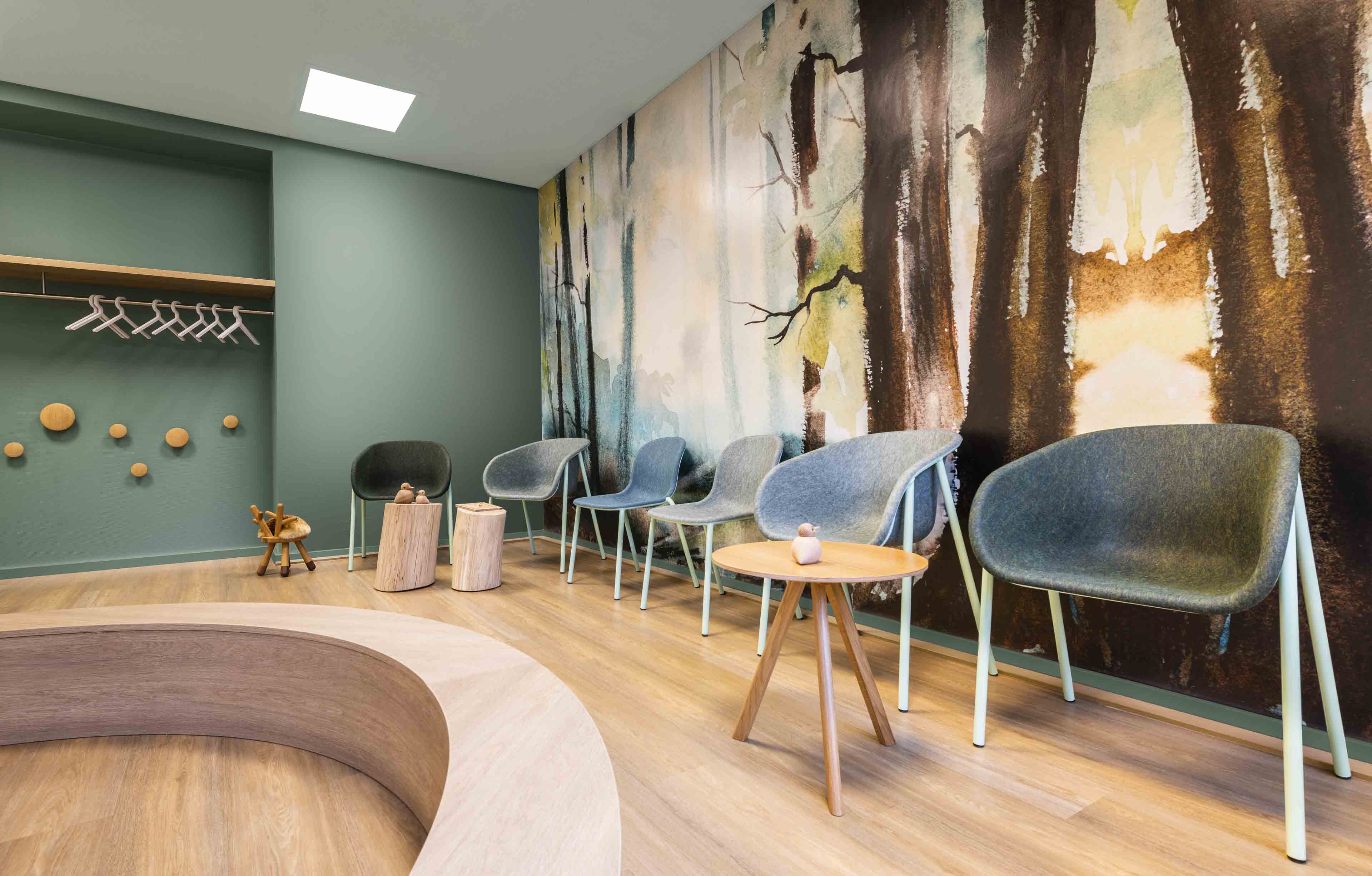 Moderner Wartebereich mit grauen Stühlen, runden Holztischen und einer Wand mit Waldlandschaft als Tapete.