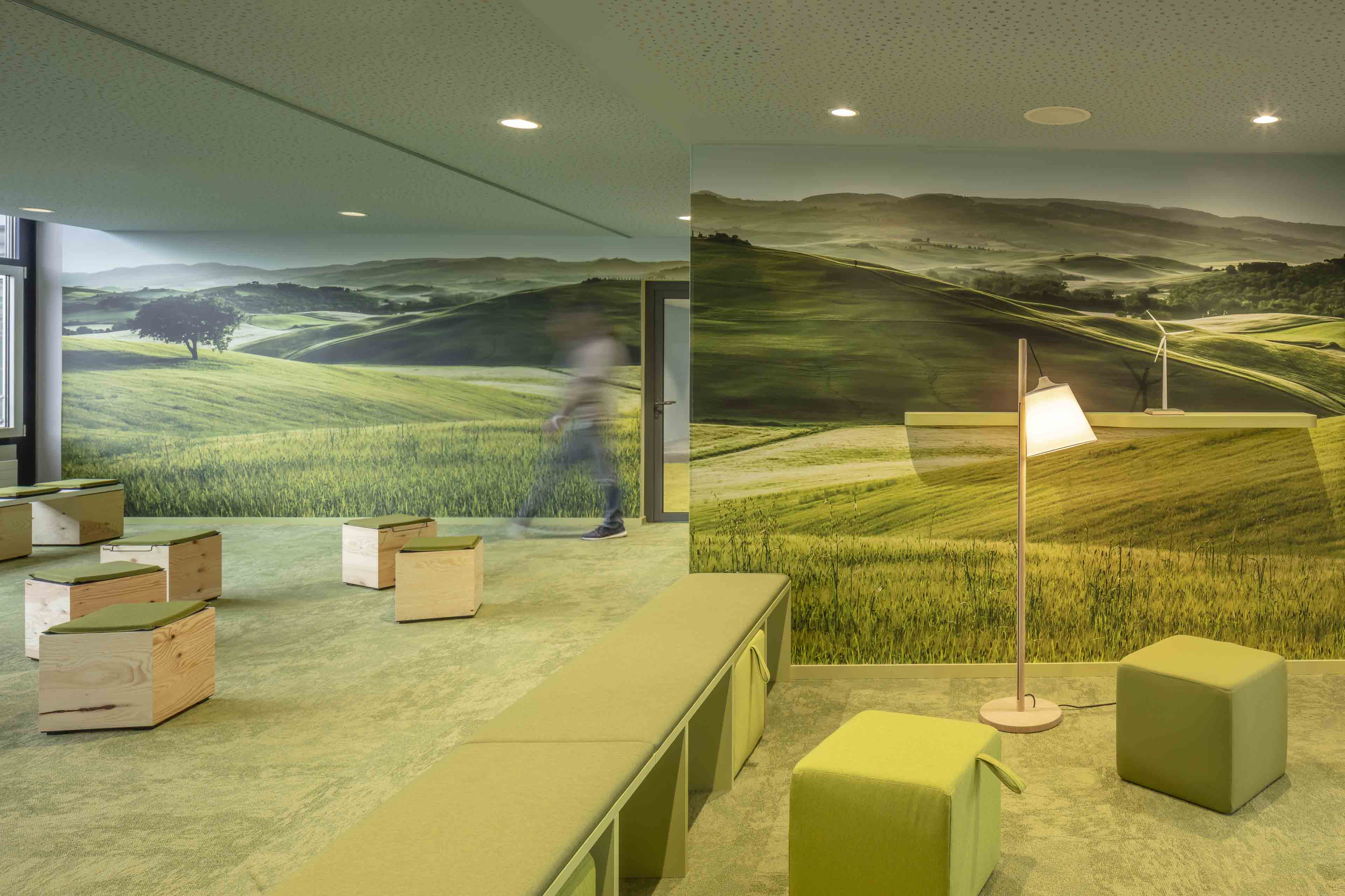 Moderner Raum mit grünen Hockern, Holzbänken und Wandbildern einer hügeligen Landschaft mit Feldern und Bäumen.