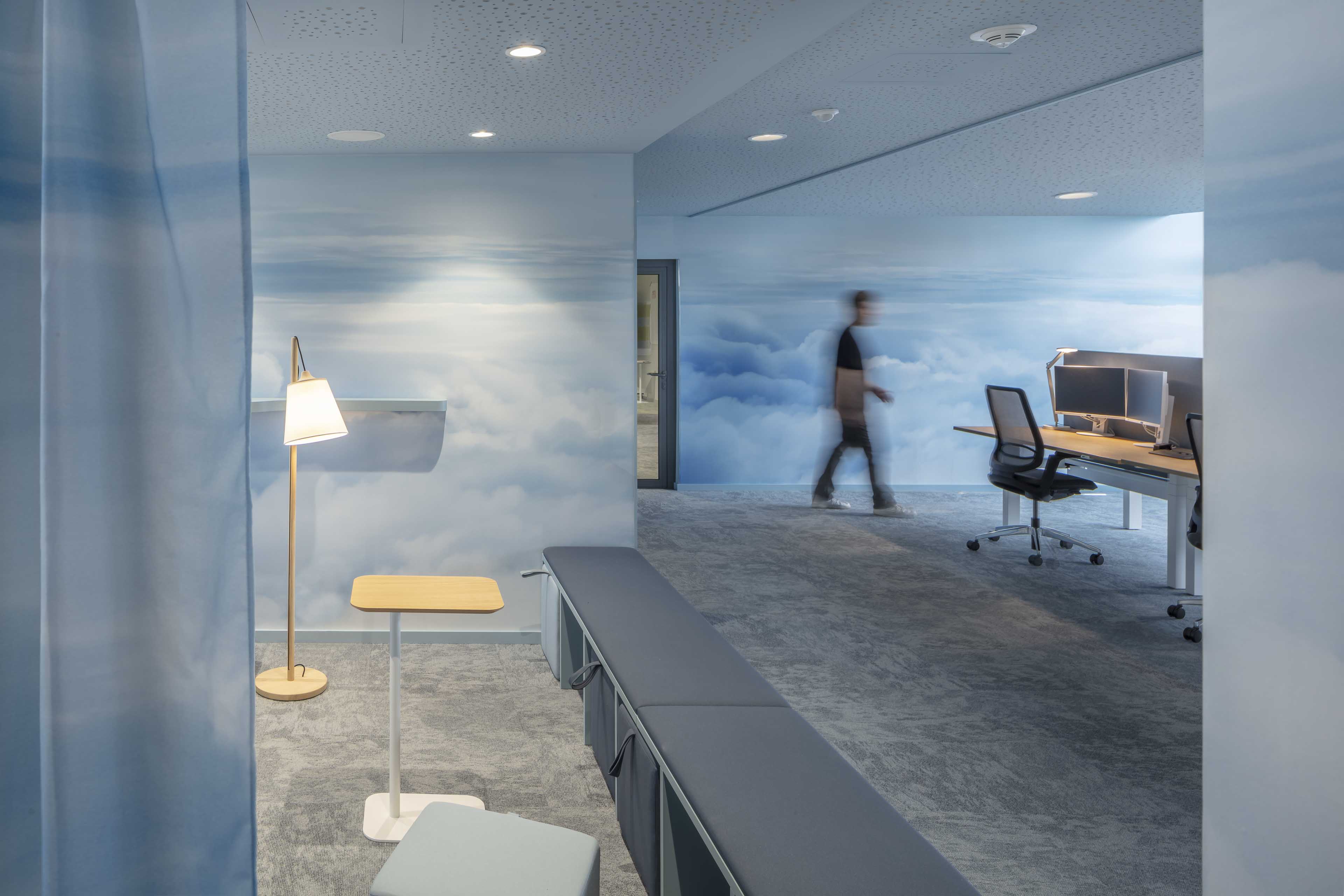 Modernes Büro mit wandfüllendem Wolkenmotiv, Schreibtischen mit Monitoren und einem verschwommenen Mann, der vorbeigeht.