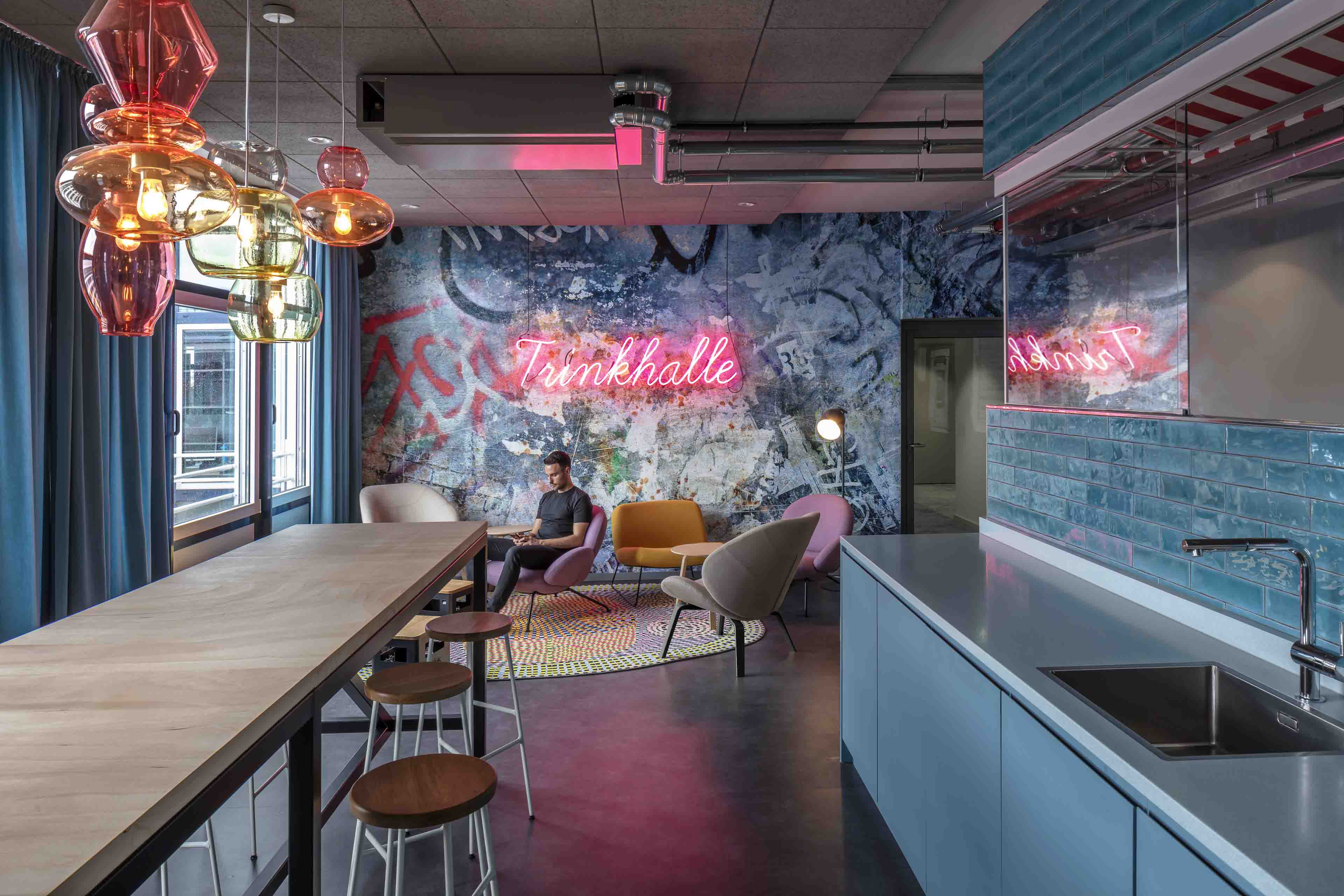 Moderne Lounge mit langem Holztisch, bunten Hängelampen, Sitzbereich mit bunten Sesseln und Neonschrift 'Trinkhalle' an der Wand.