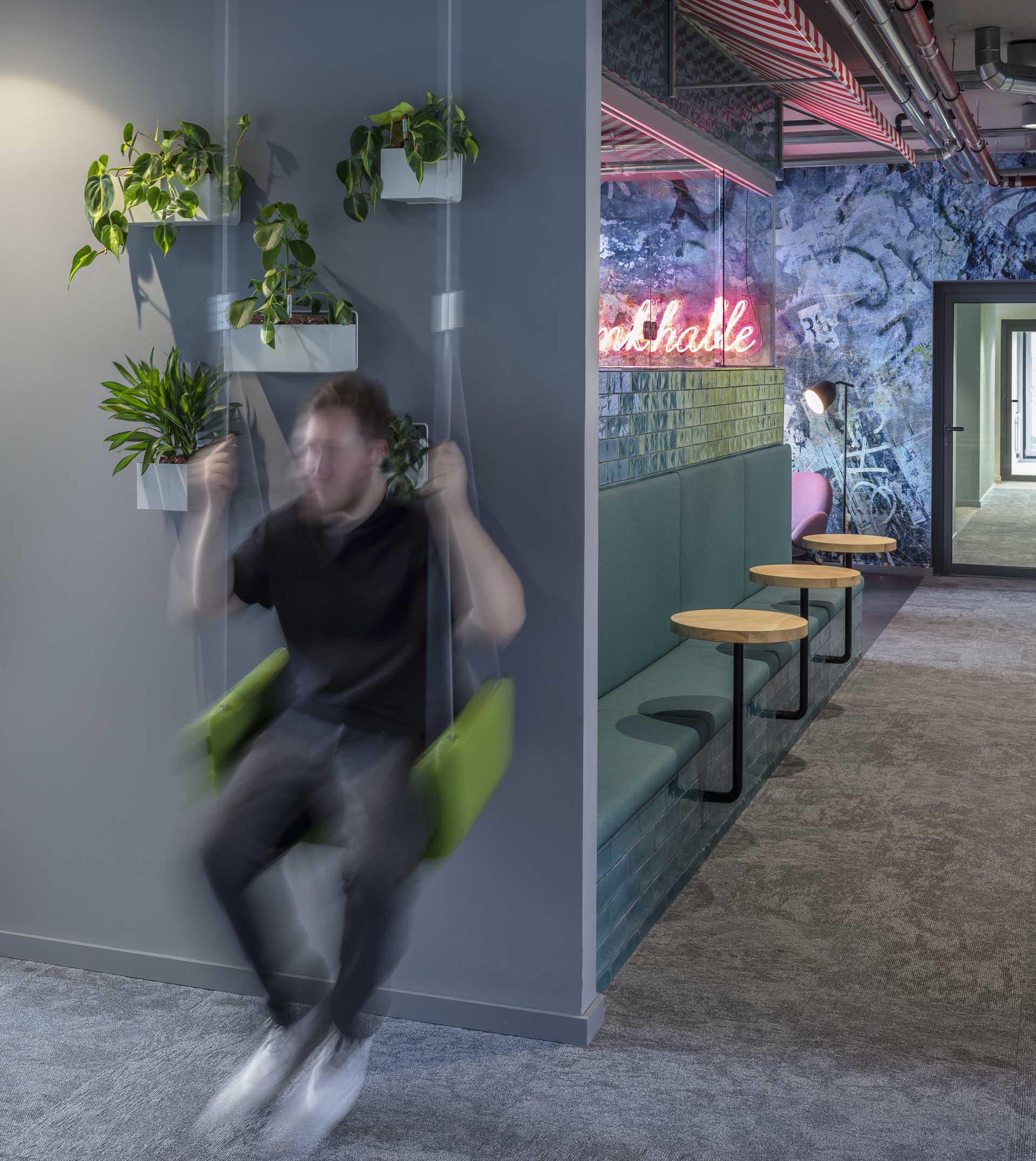 Unscharfe Aufnahme eines Mannes, der in einem modernen Innenraum an einer grünen Schaukel schwingt, mit Wandpflanzen links und einer Sitzbank mit Tischen rechts.