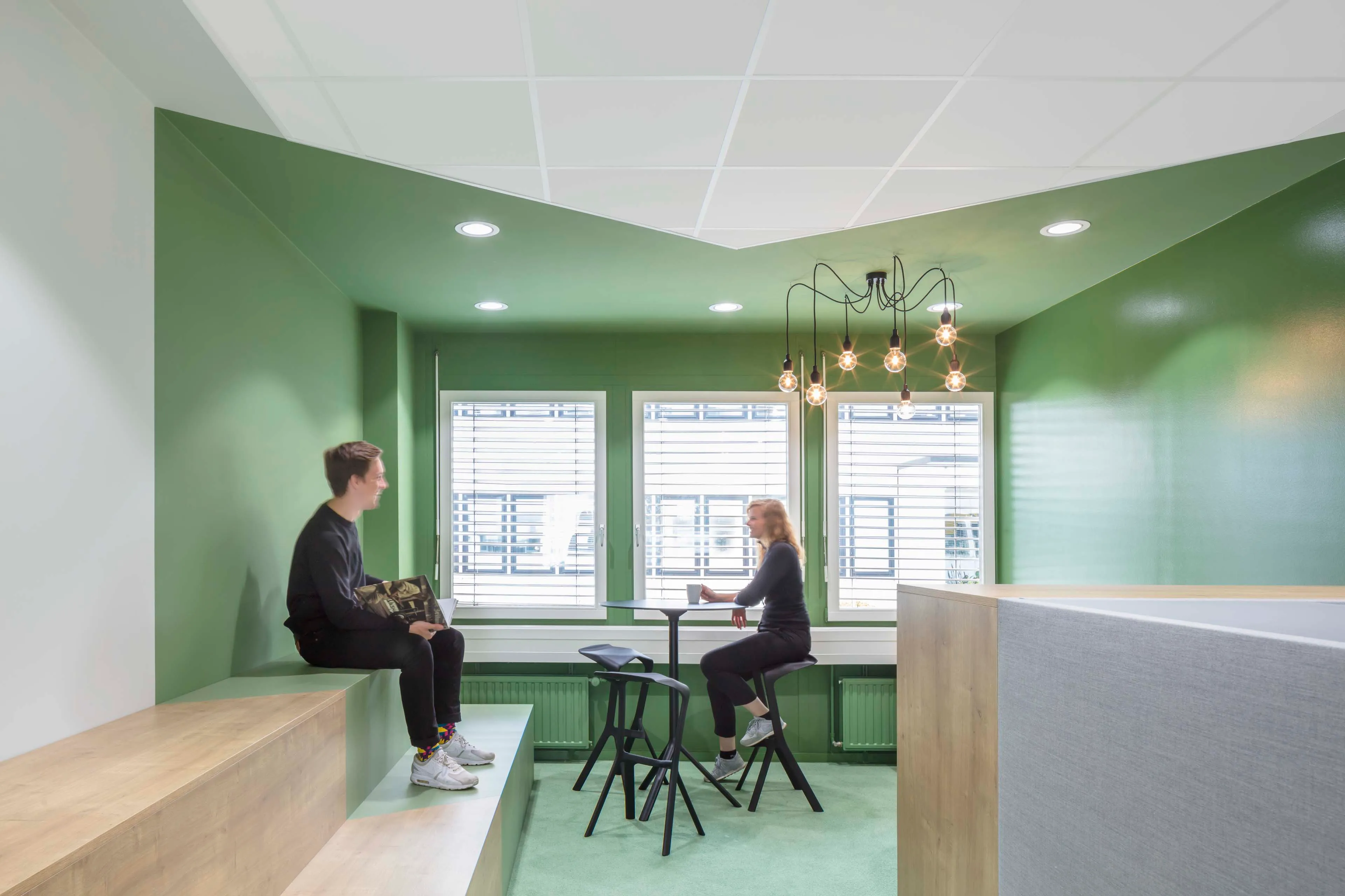 Zwei Personen unterhalten sich in einem modernen, hellgrünen Raum mit großen Fenstern, Stehtisch und Hockern.