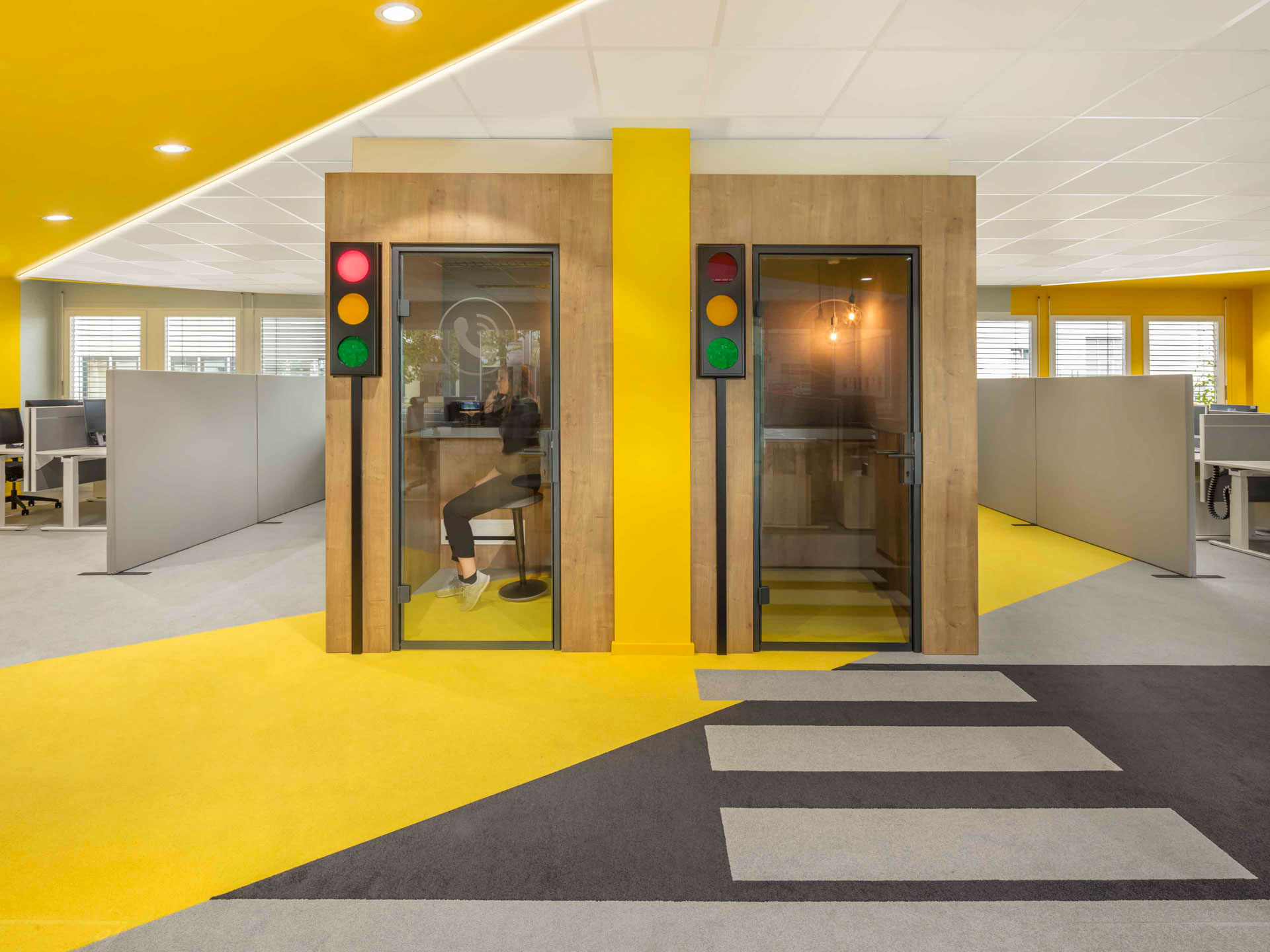 Moderne Bürofläche mit zwei schallisolierten Telefonkabinen, eine links mit einer sitzenden Person, Gelb- und Grautönen auf dem Boden und einer Fußgängerampel zwischen den Kabinen.