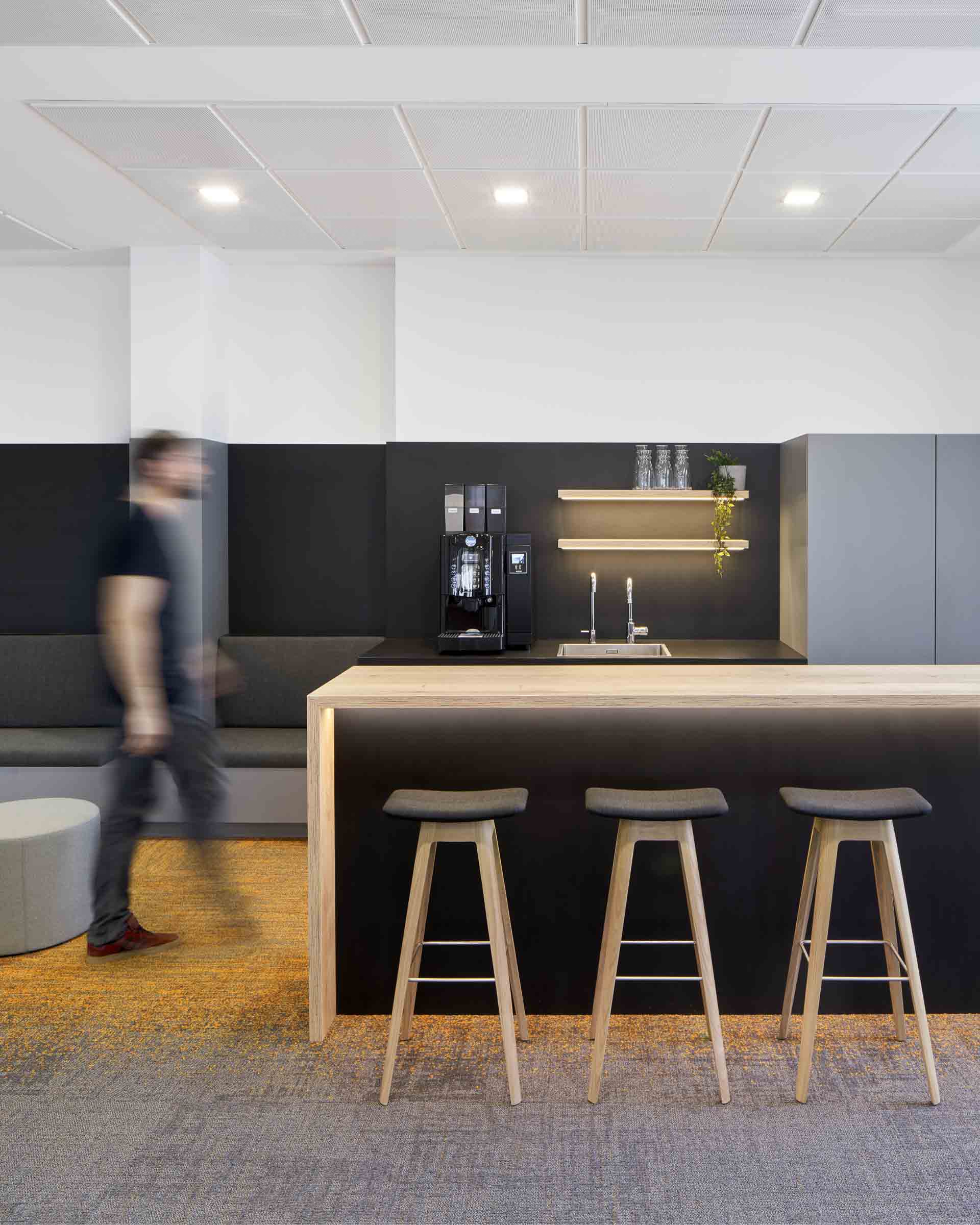 Moderne Küche in einem Büro mit drei Holzbarhockern, Kaffeeautomat und zwei Regalen mit Gläsern und Pflanze.