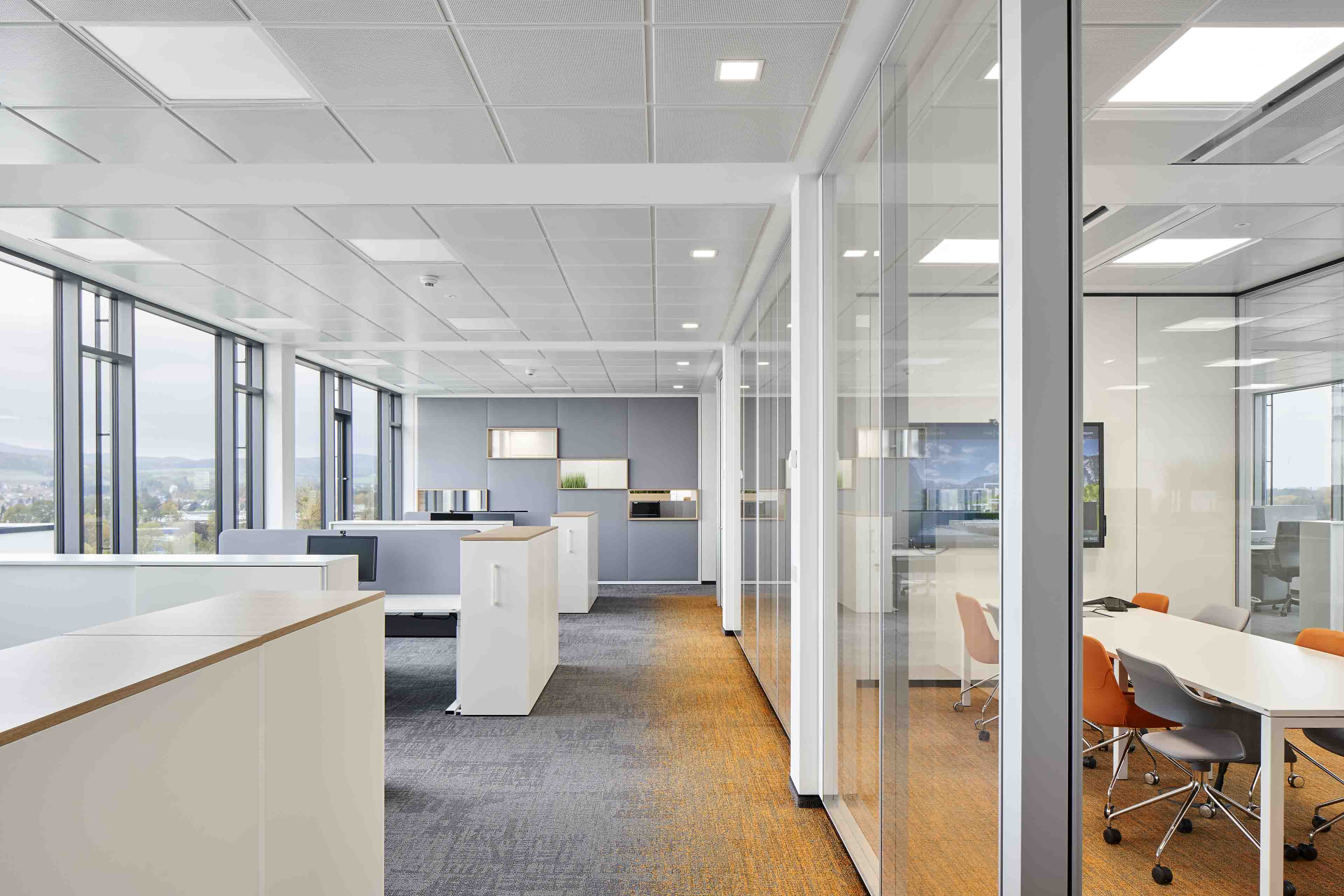 Modernes helles Büro mit Glaswänden, mehreren Schreibtischen und bunten Stühlen in einem Besprechungsraum.