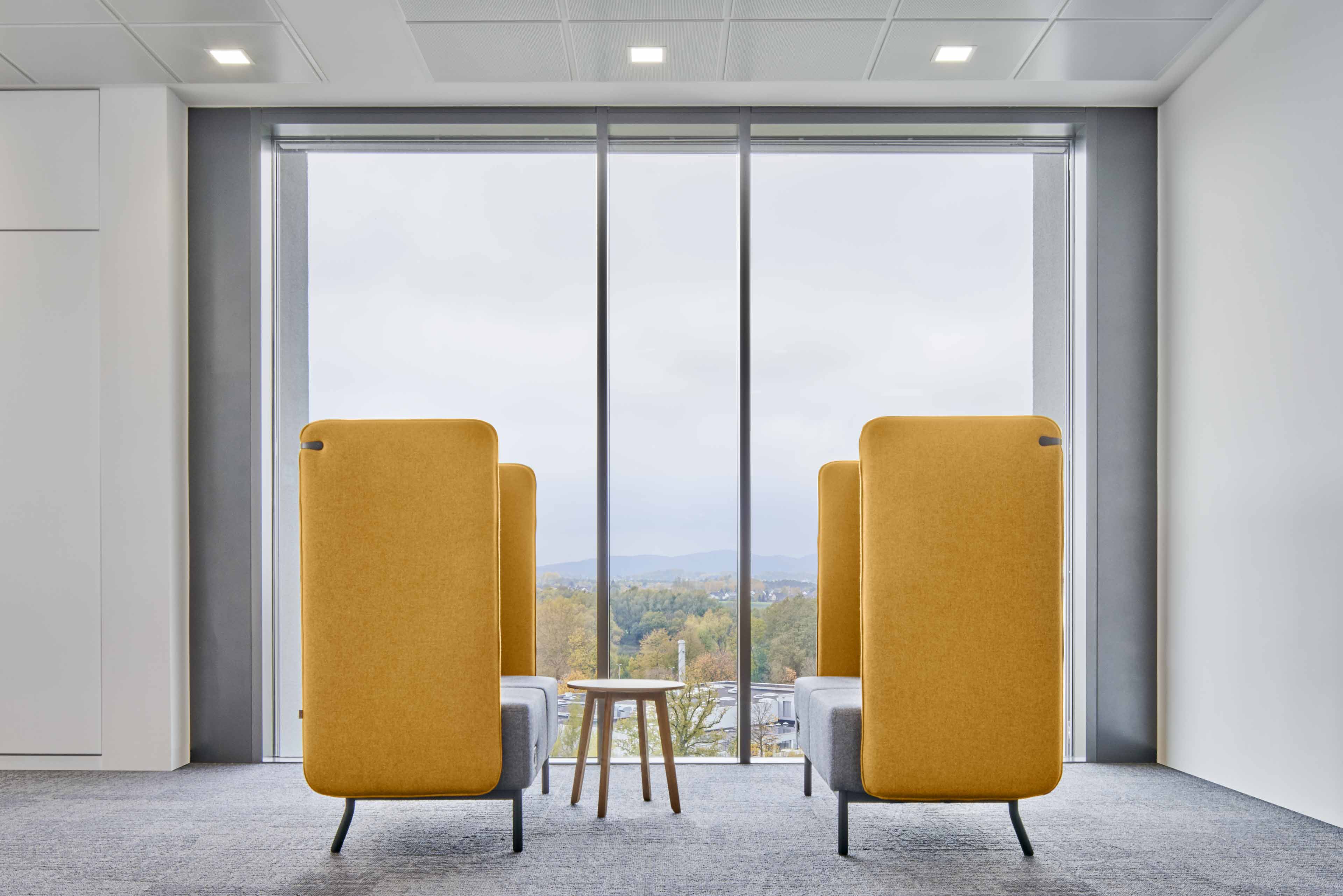 Moderner Sitzbereich mit orangefarbenen Sofas und kleinem Holztisch vor großer Fensterfront mit Blick auf Bäume und Hügel.