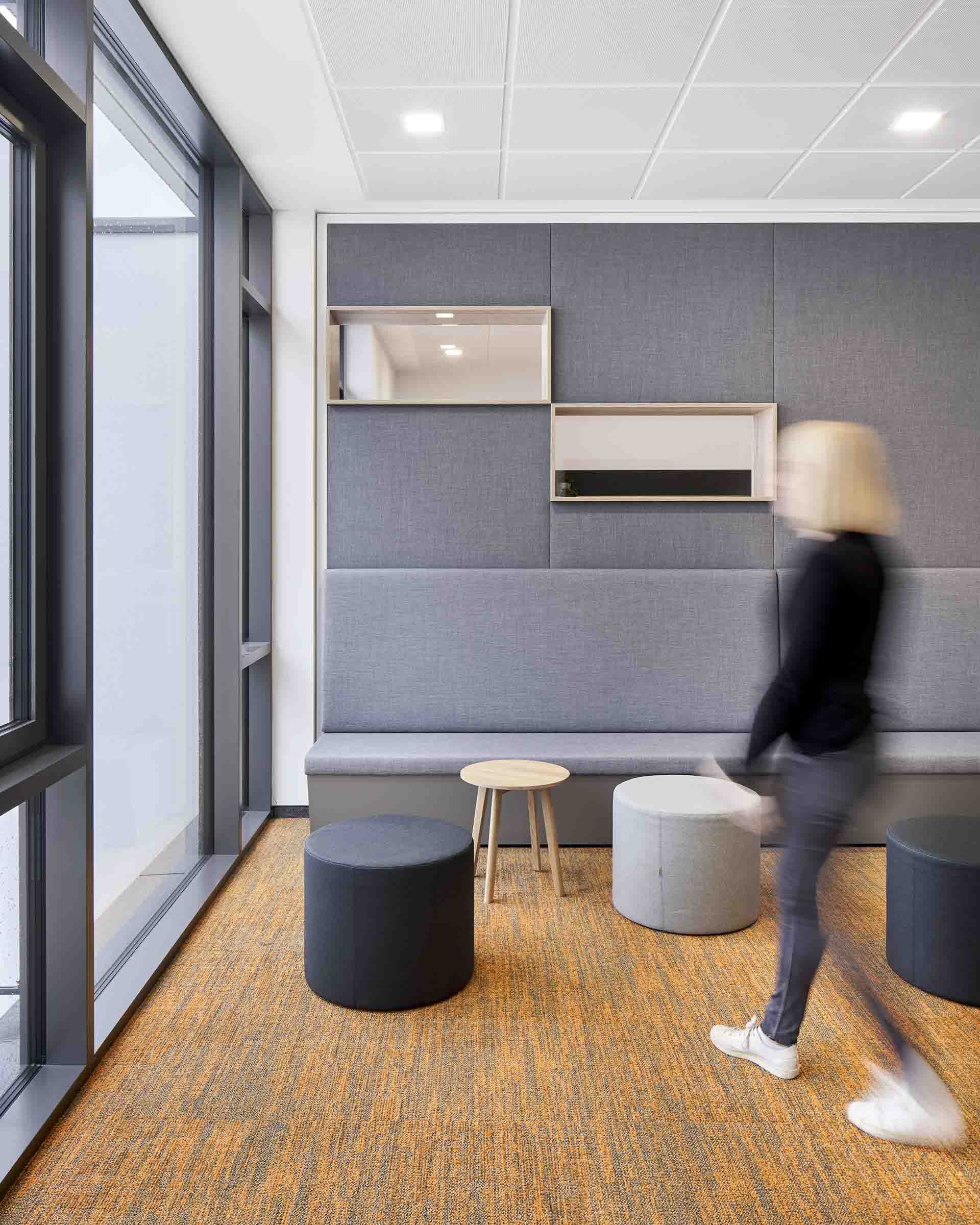 Moderne Lounge mit grauer Polstersitzbank, runden Hockern, kleinem Holztisch und einer streichenden Person mit blonden Haaren.