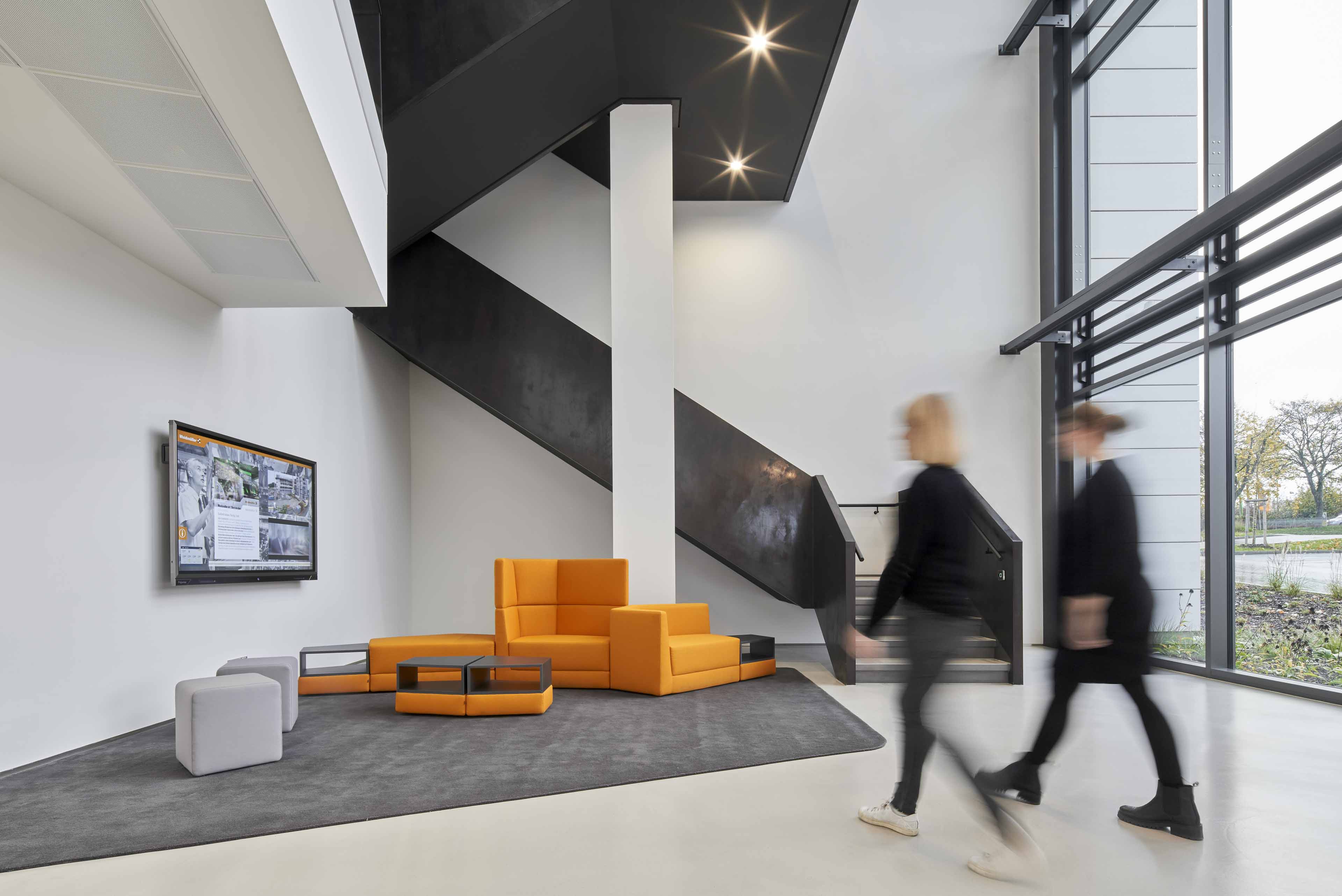 Moderner Wartebereich mit orangen und grauen Sitzmöbeln, schwarzer Treppe und großen Fenstern mit Blick nach draußen.