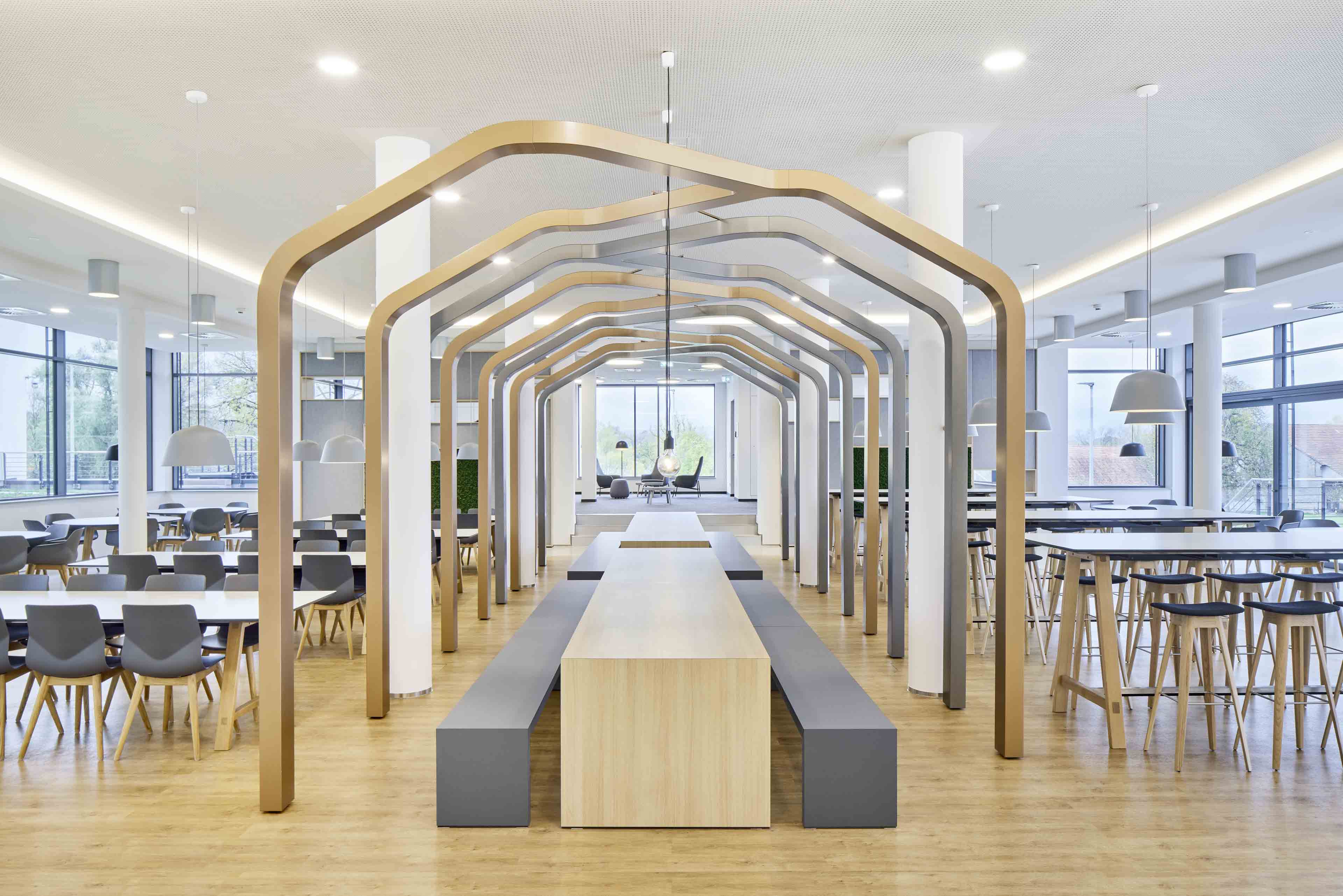 Heller moderner Essbereich mit langen Holztischen, grauen Bänken, Stühlen und dekorativen Rahmen aus Holz und Metall.