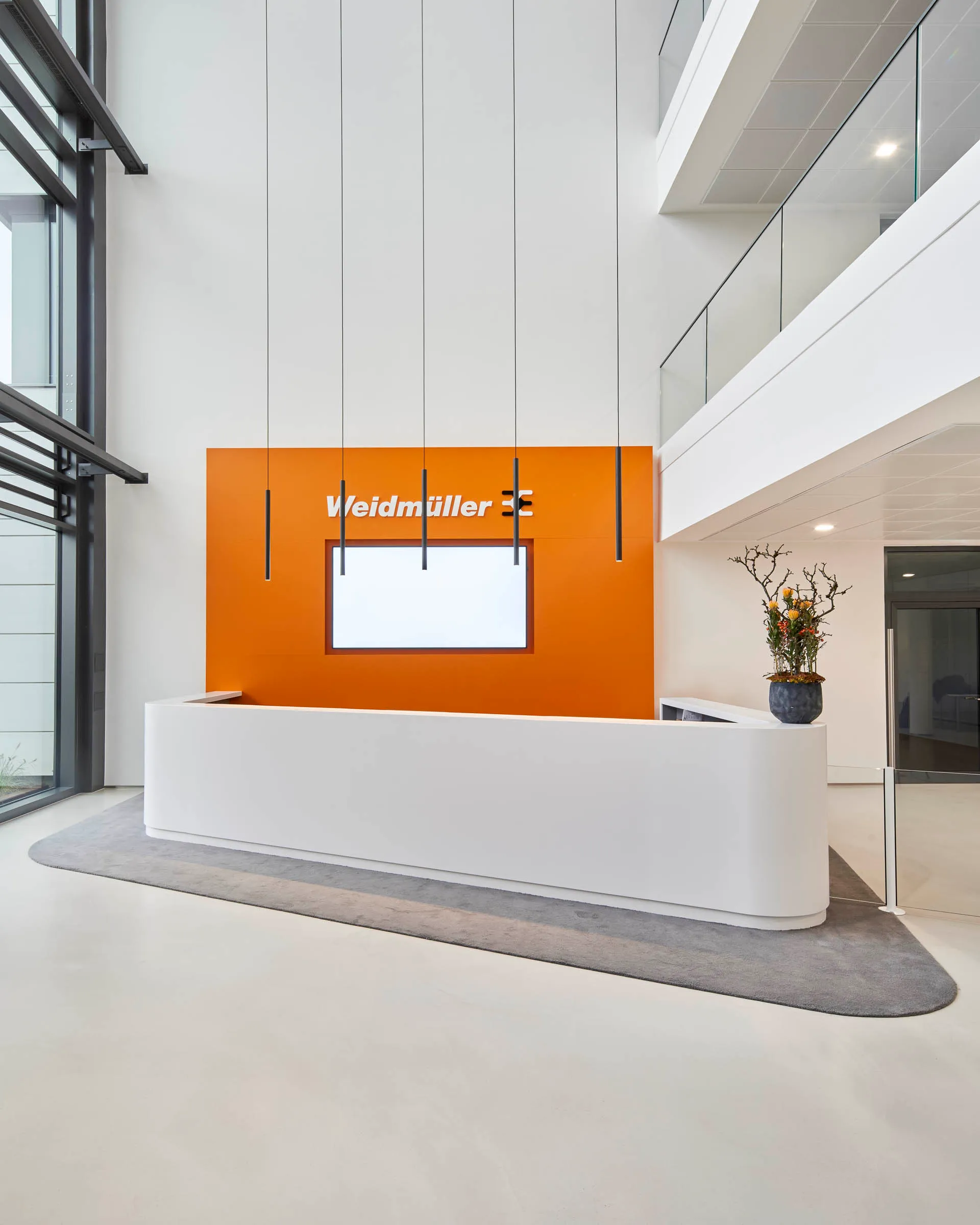 Moderner Empfangsbereich mit weißem Tresen vor orangefarbener Wand mit Weidmüller-Logo und hängenden schwarzen Pendelleuchten.