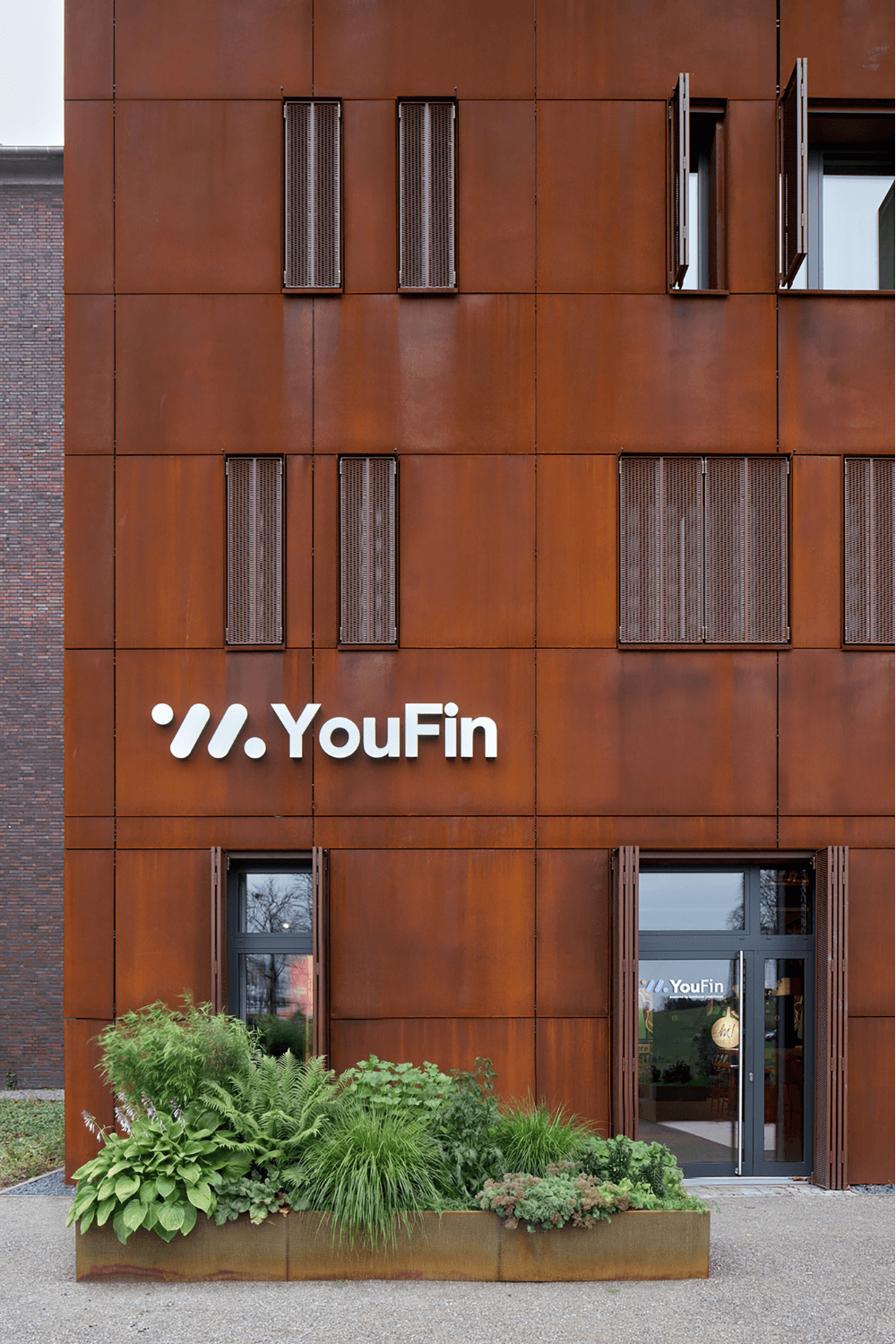 Gebäudefassade aus rostbraunen Metallplatten mit dem weißen Schriftzug 'YouFin' und einem begrünten Pflanzbeet davor.