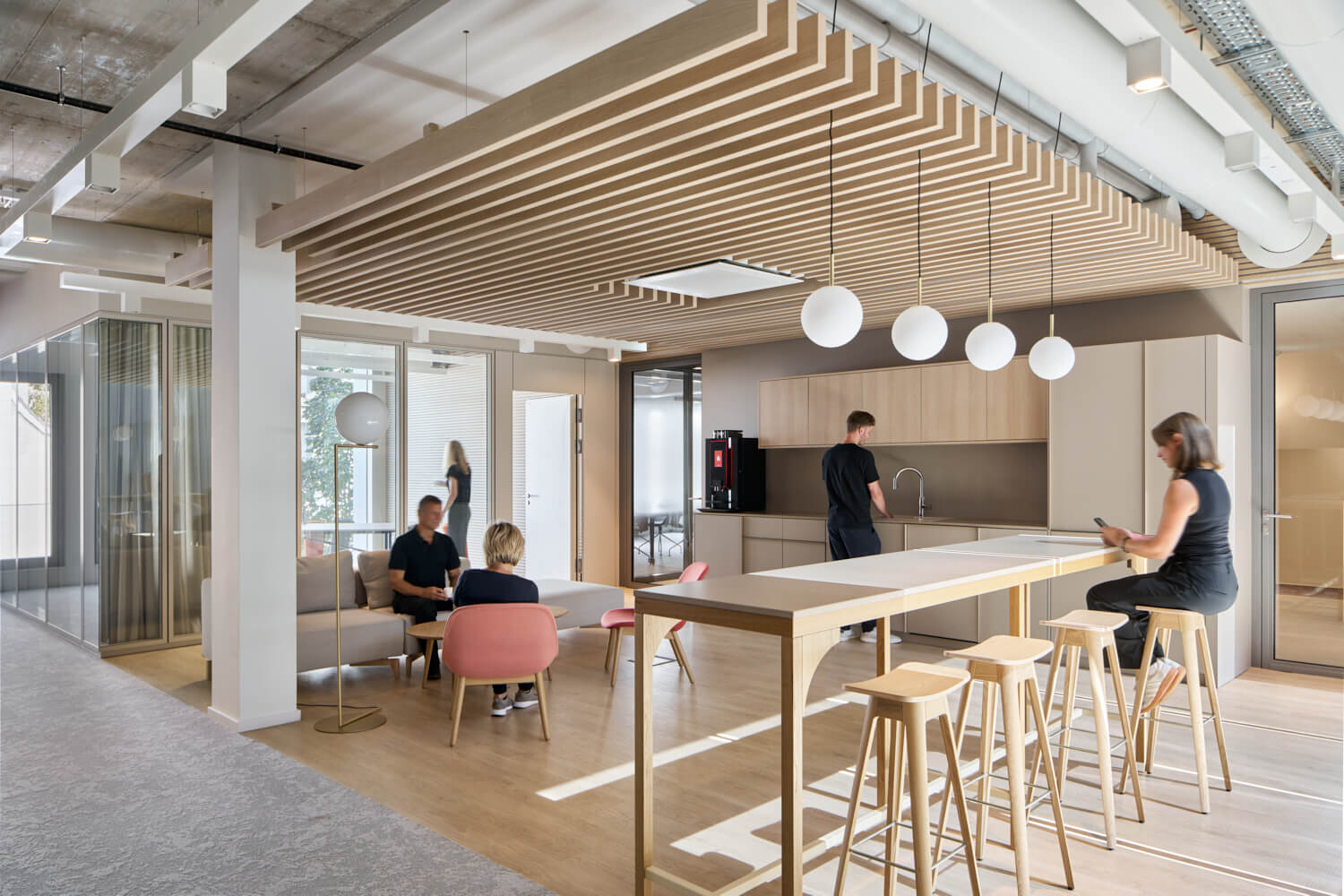 Moderner Büro-Küchenbereich mit Holztisch, Hockern, mehreren Personen und geometrischer Deckendekoration.